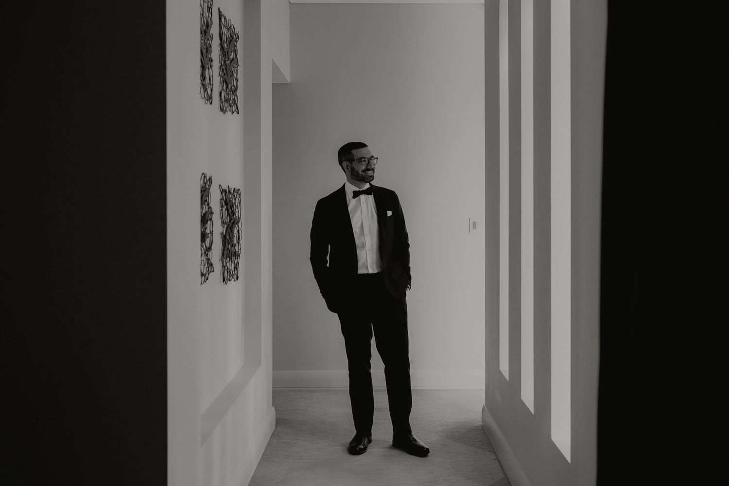 Groom in black tuxedo standing in elegant hallway with artwork, black and white wedding portrait.