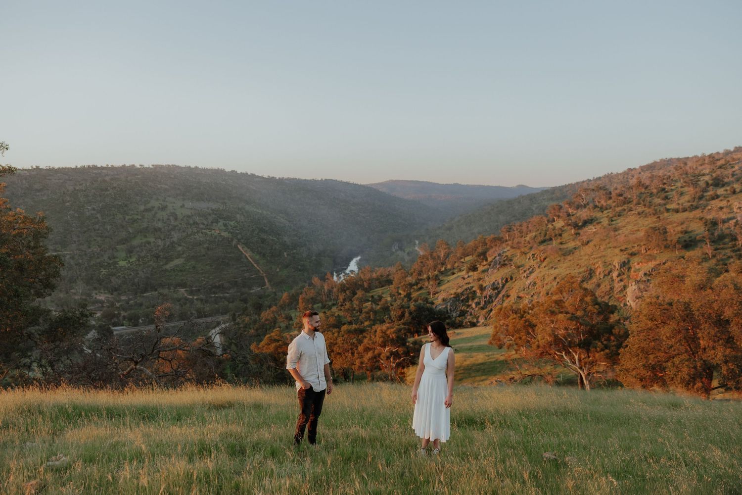 Couple in white outfits standing in green meadow with stunning mountain valley backdrop at dusk.