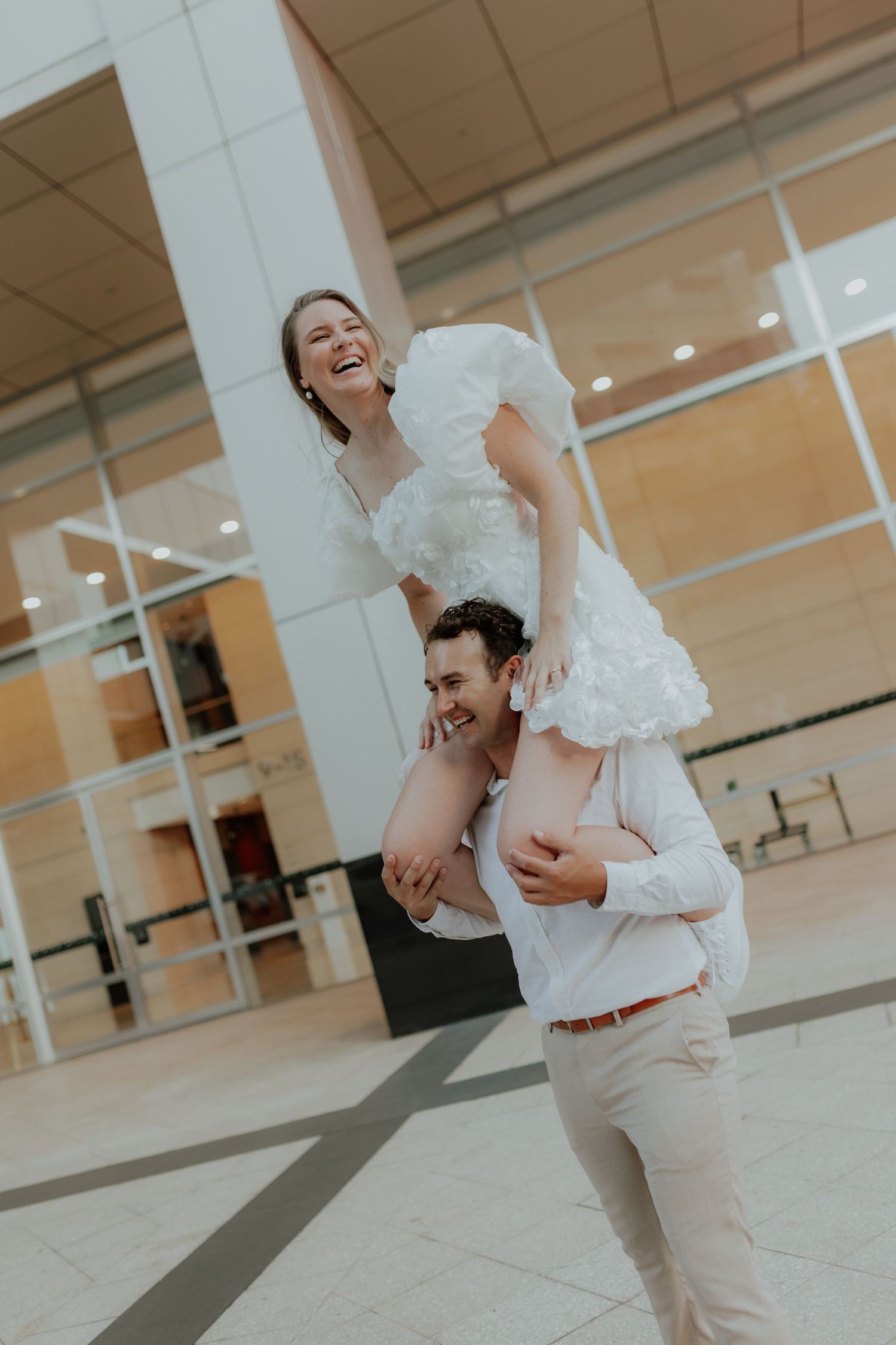 Groom lifting bride on his shoulders as she laughs joyfully in a white ruffled wedding dress outdoors.