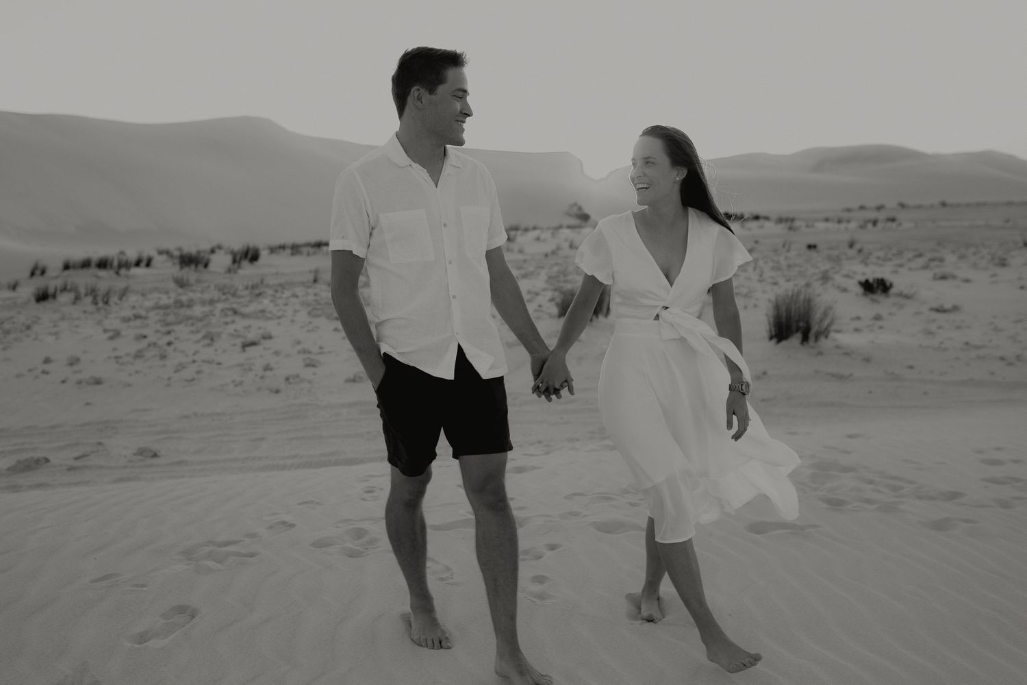 Black and white photo of a couple holding hands and walking barefoot on sand dunes in a desert landscape.