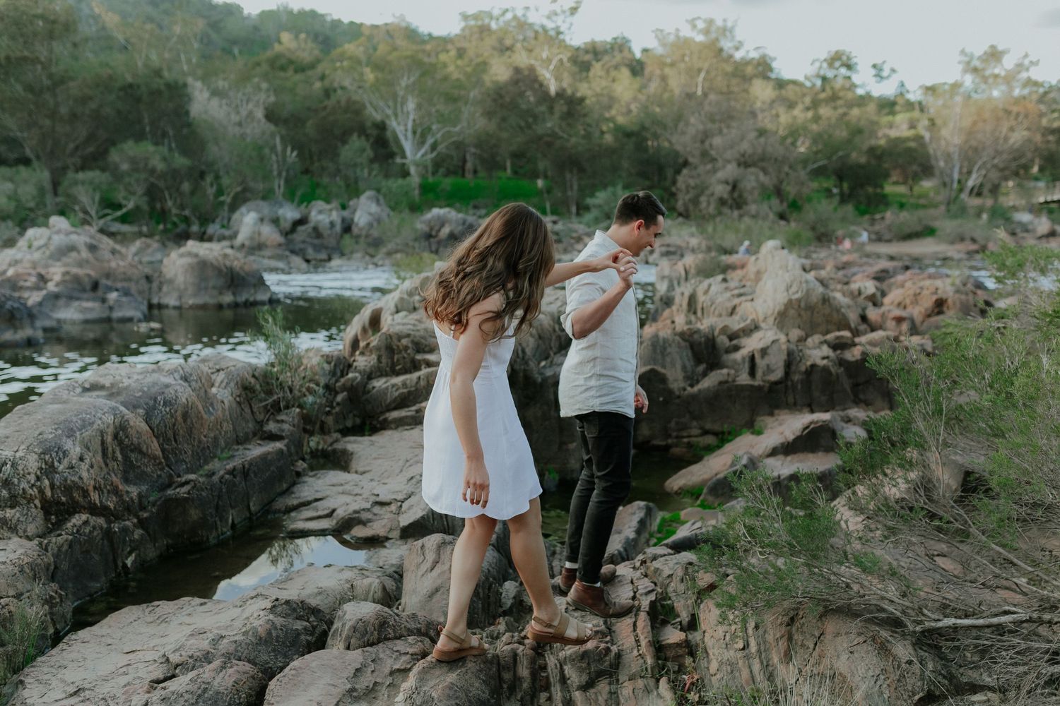 Couple holding hands while carefully navigating rocky terrain near a river surrounded by lush green trees.