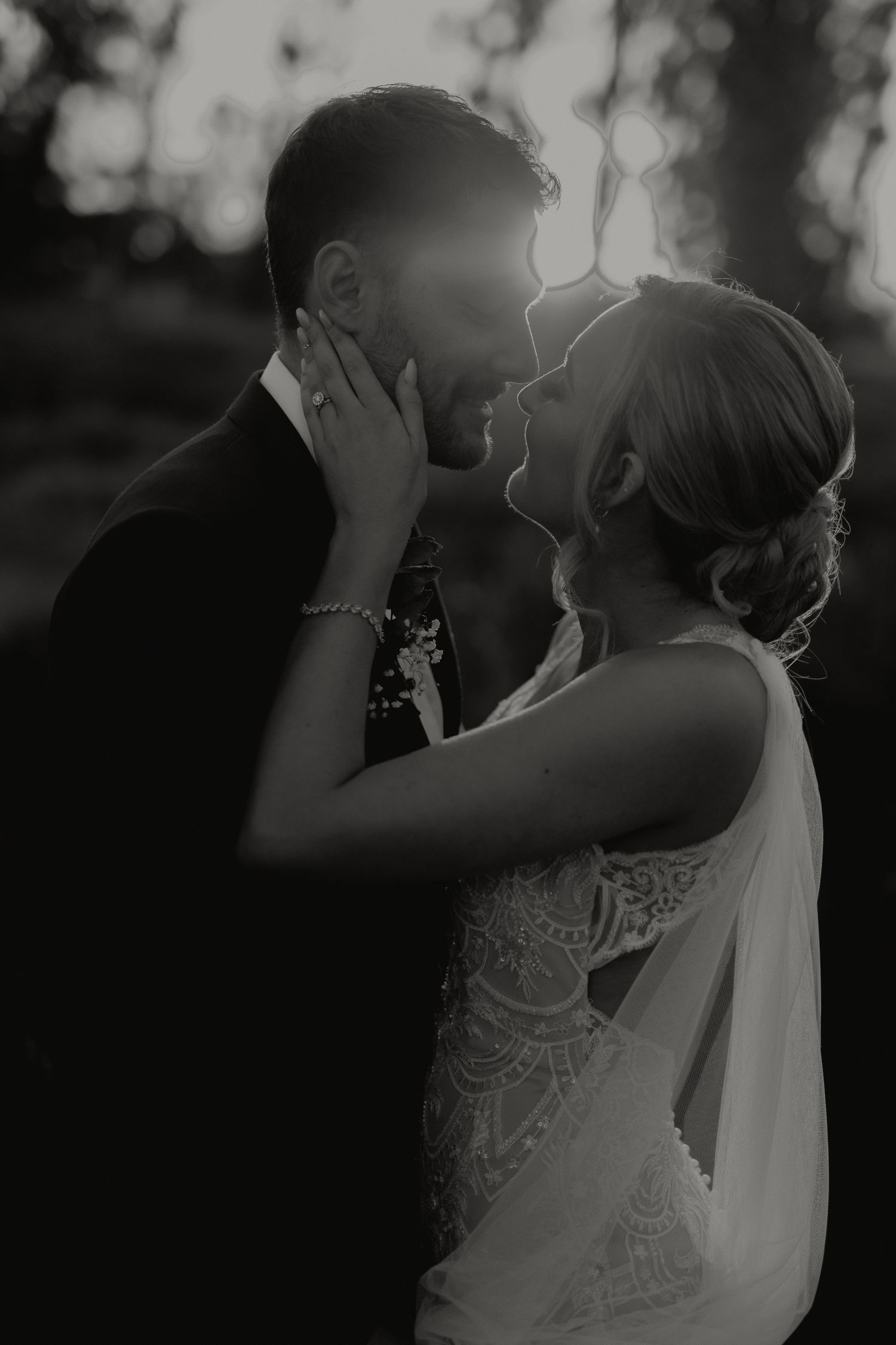 Black and white romantic photo of bride and groom almost kissing at sunset, backlit by golden light.