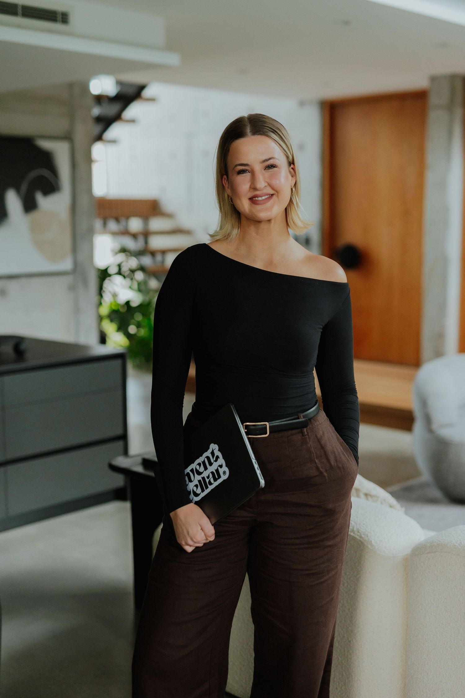 Blonde woman in black off-shoulder top and brown wide-leg pants holding an Event Cellar notebook.