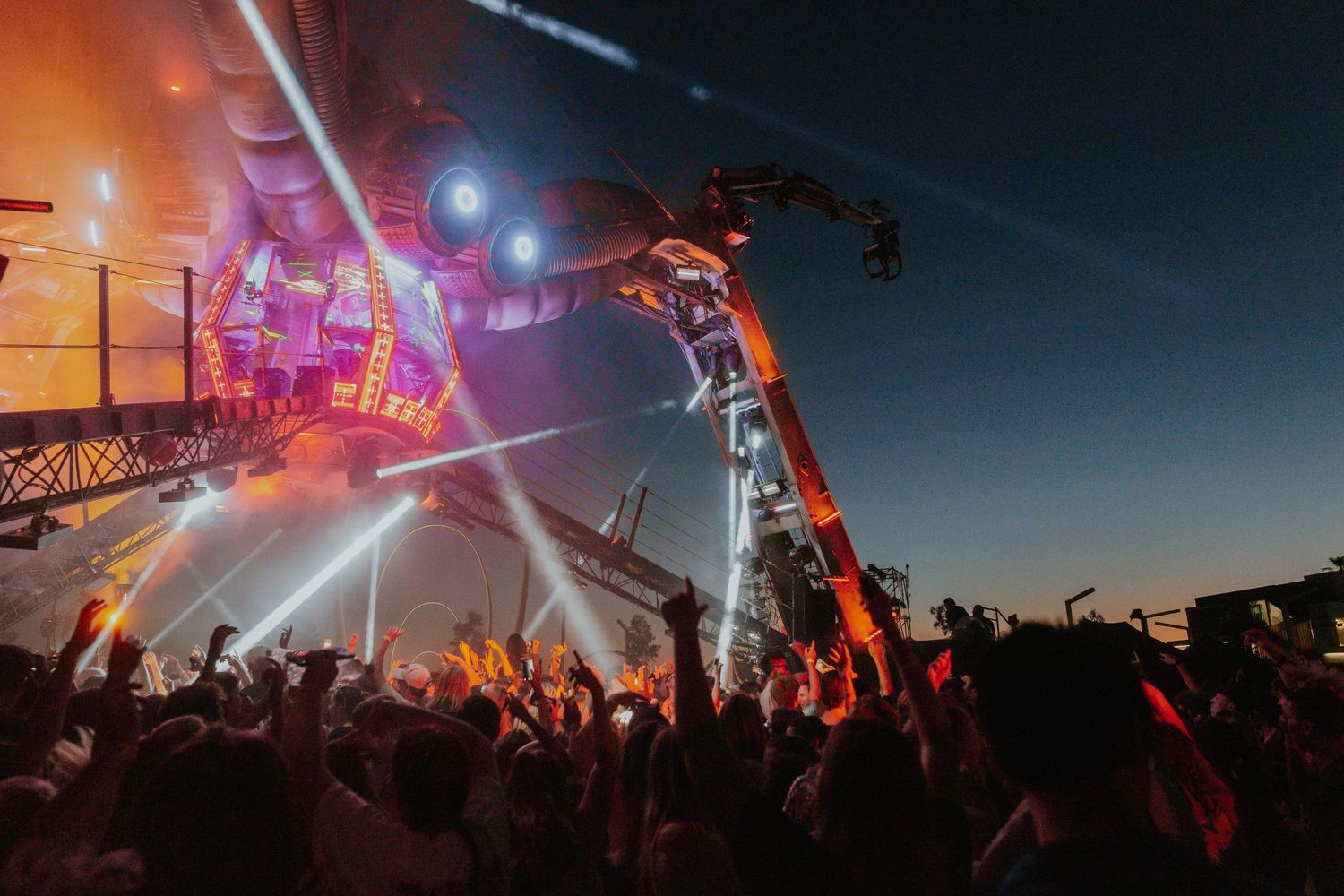 Energetic music festival crowd raises hands under spectacular stage lighting at twilight outdoor concert.