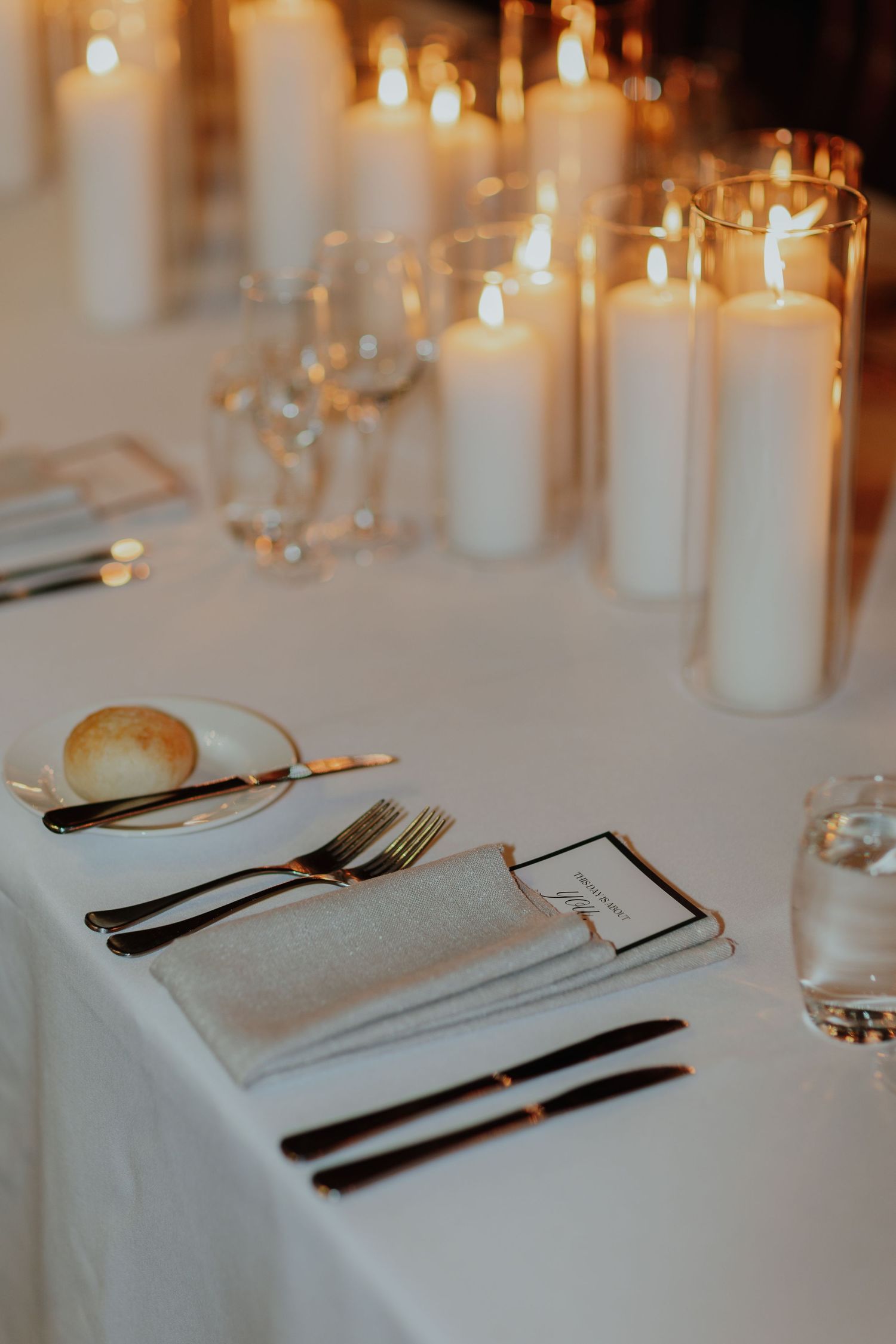 Elegant place setting with gold cutlery, white napkin, and glowing white pillar candles on table.