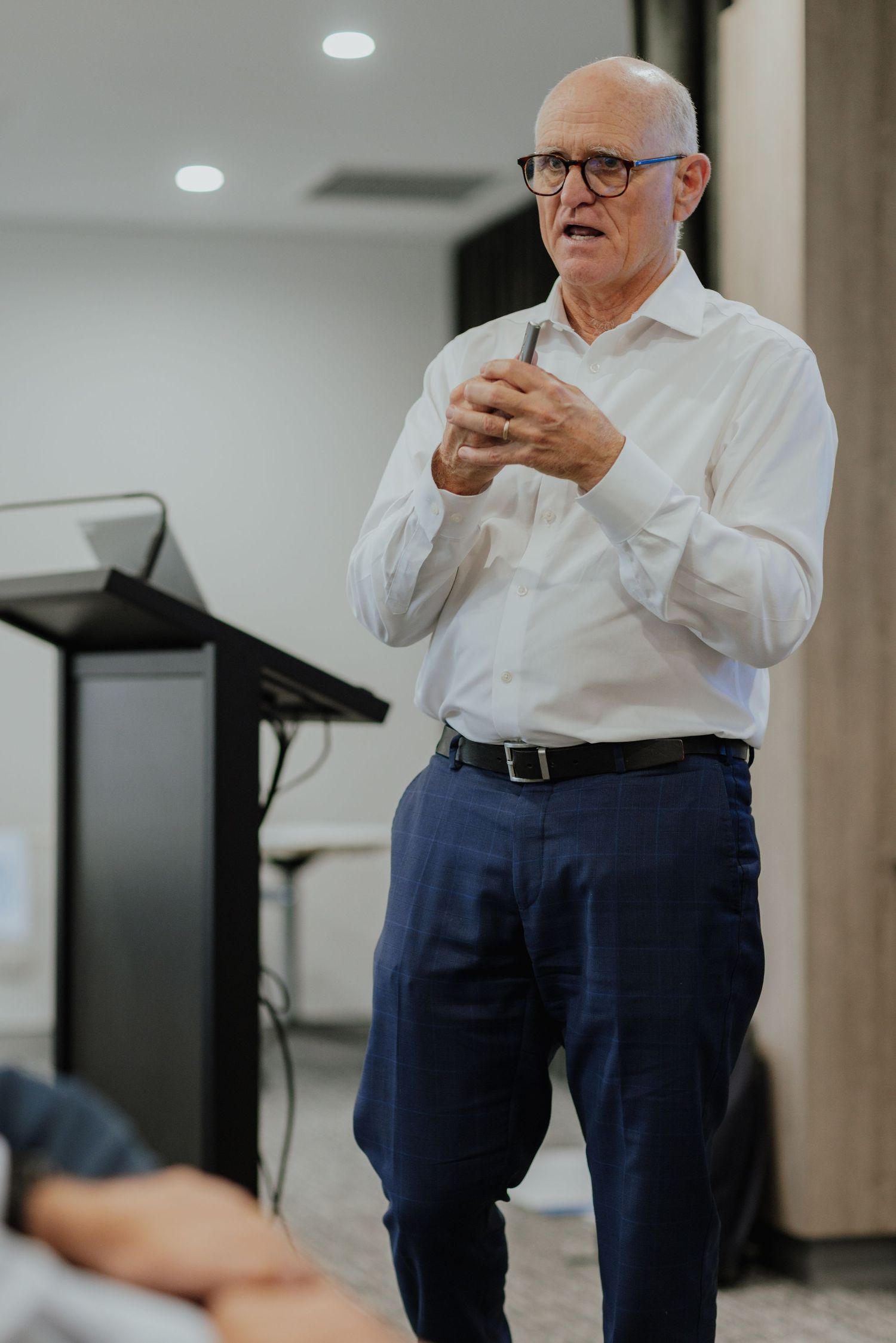 An older businessman in white shirt and blue pants speaks confidently during a corporate presentation.