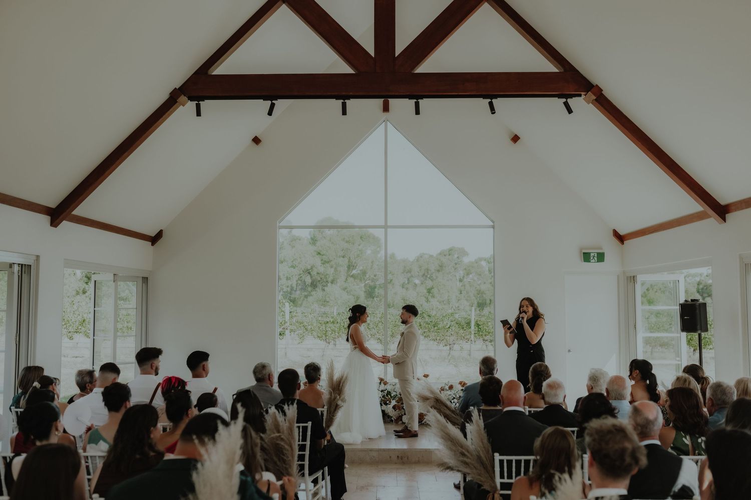 Wedding ceremony inside a white chapel with vaulted timber ceilings and large triangular window.