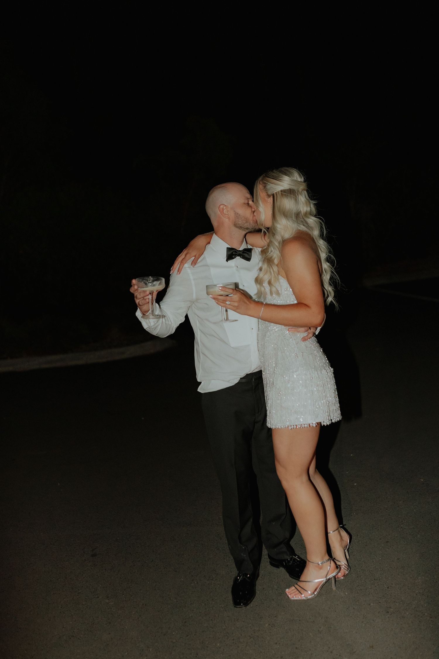 Bride and groom kissing at night holding cocktail glasses, bride wearing a sequined mini dress.