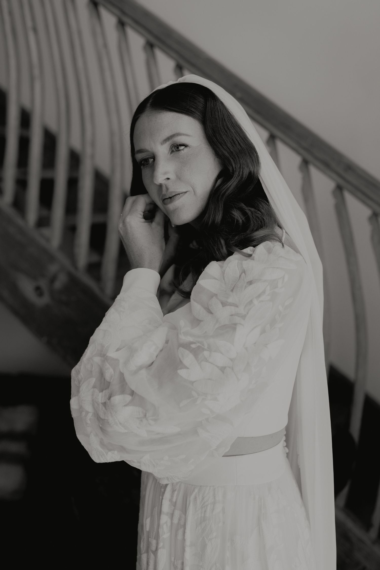 Elegant bride in a textured white gown with puff sleeves and veil posing on a staircase, black and white.