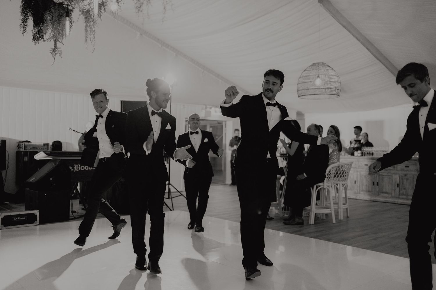 Black and white photo of groomsmen in tuxedos dancing energetically on white dance floor at wedding reception.