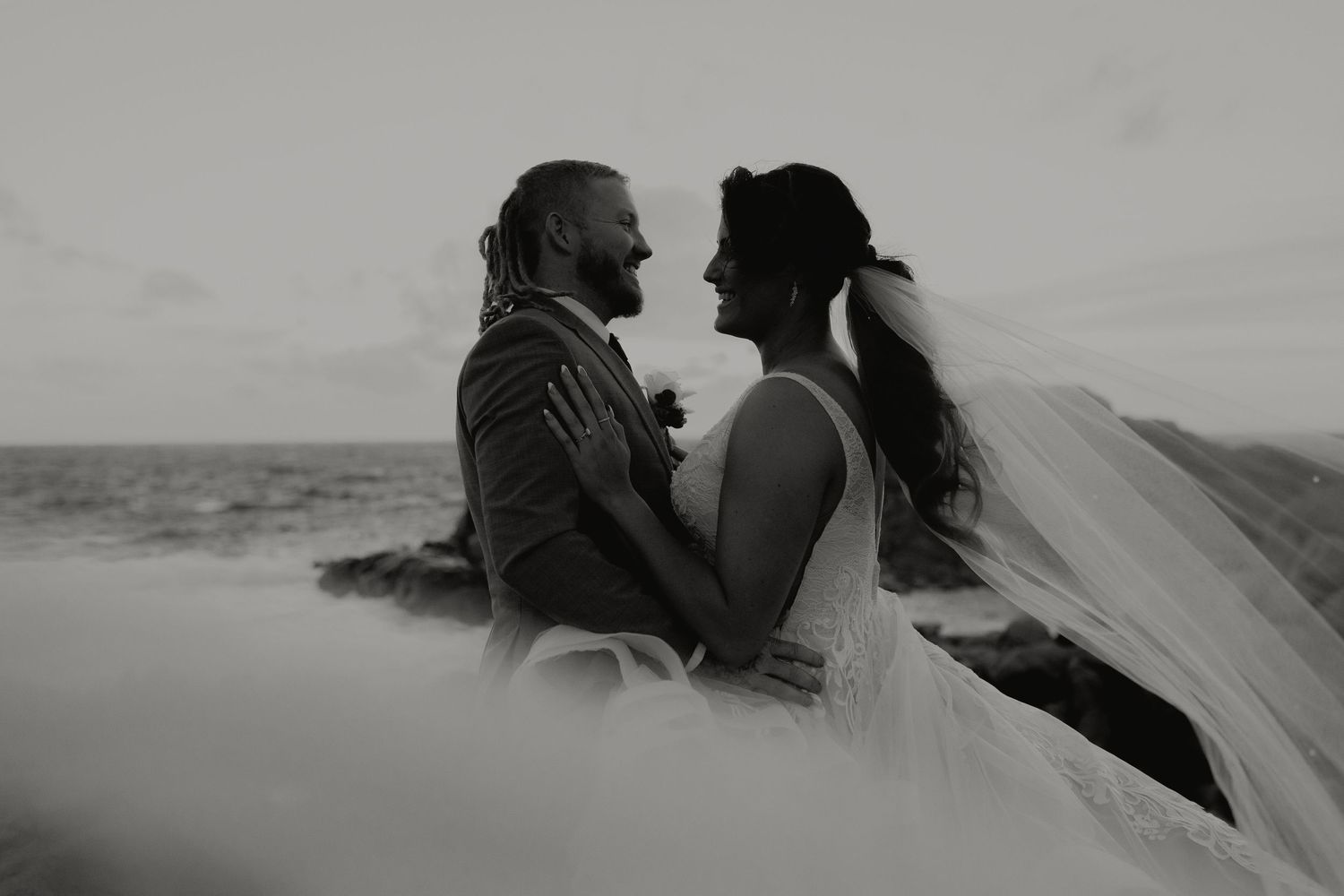 Black and white photo of bride and groom embracing by the ocean.