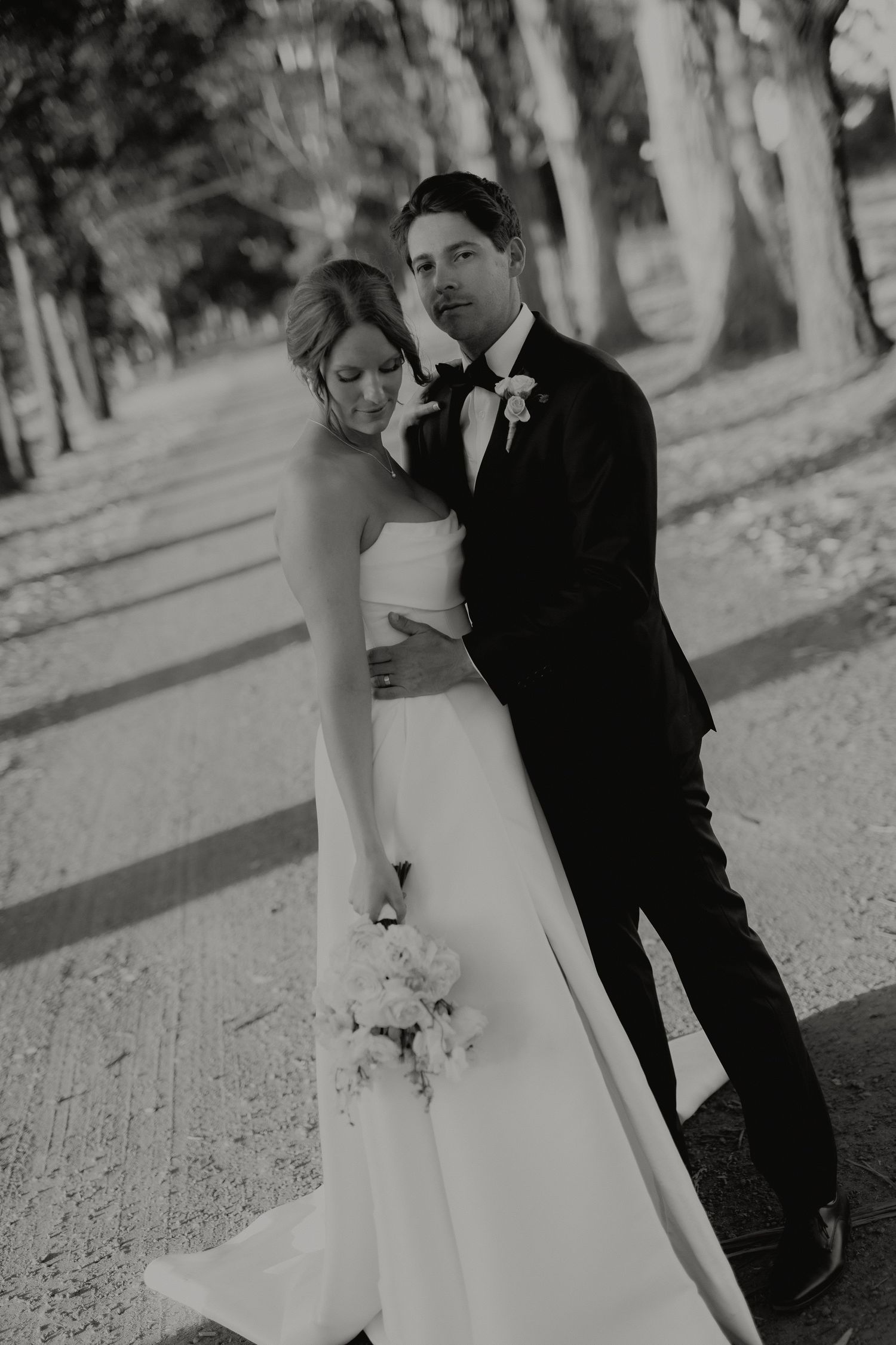Black and white portrait of bride leaning on groom in tuxedo along tree-lined path.