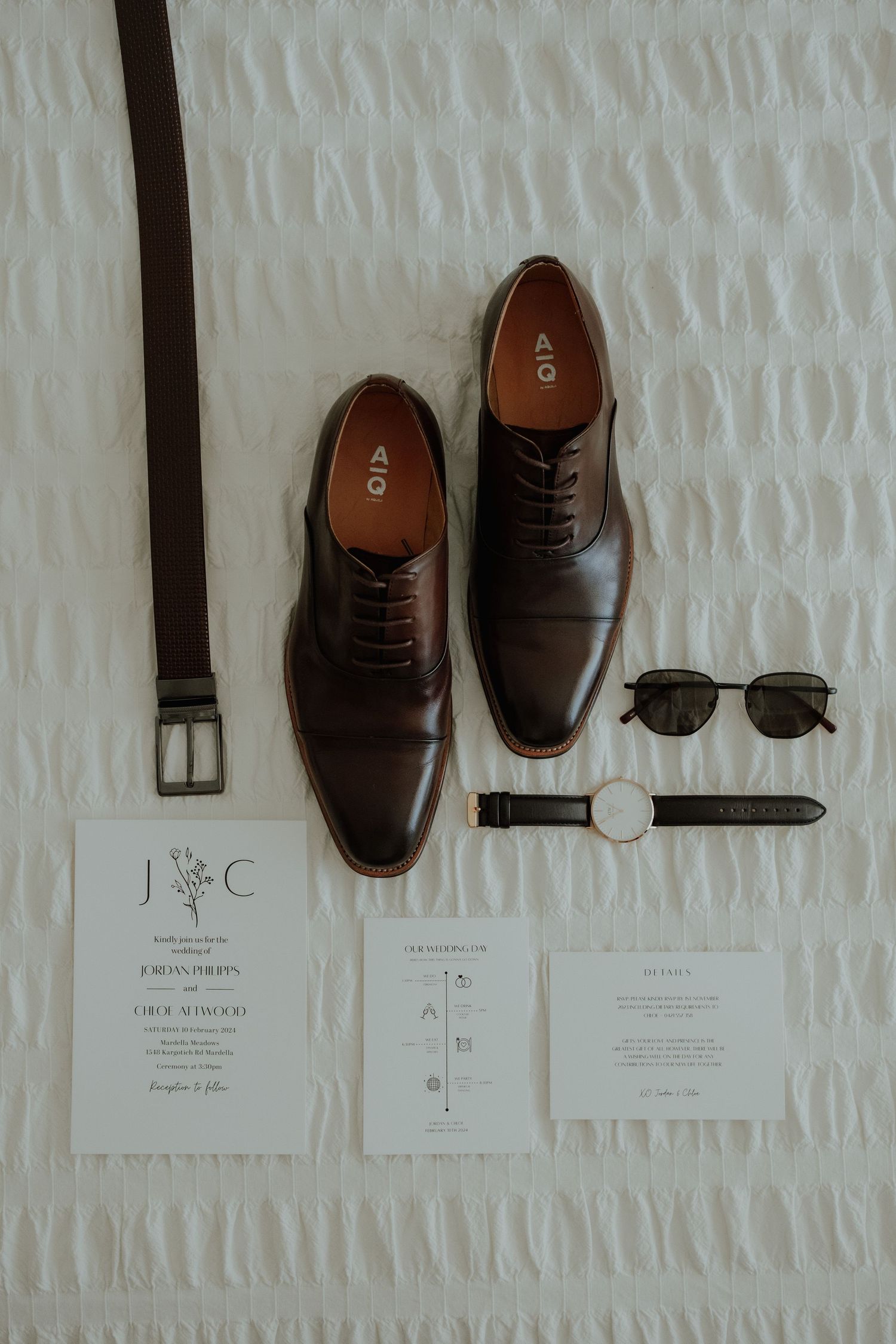 Brown leather Oxford shoes with matching belt, watch, sunglasses, and wedding invitation flatlay on white textured surface.