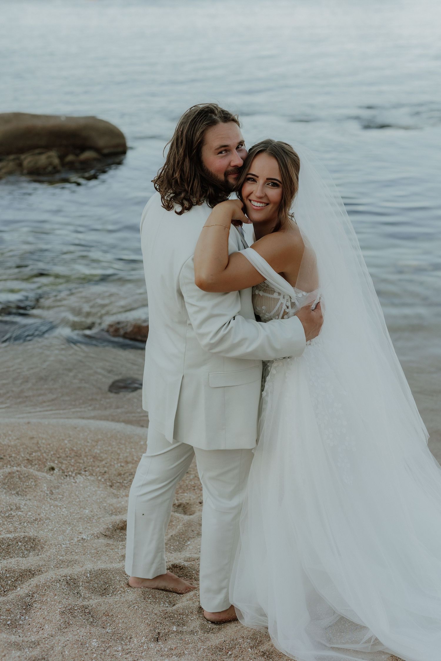 Smiling bride and groom embracing on sandy beach with ocean rocks in the background.