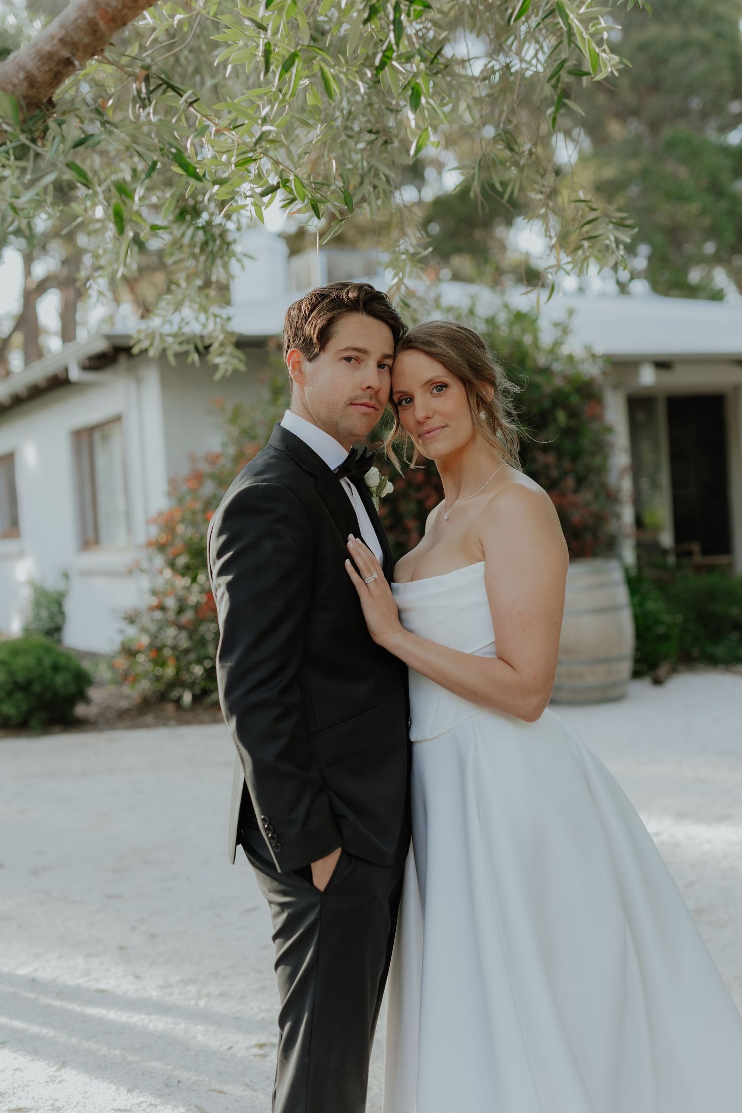 Bride and groom embracing tenderly outdoors near olive tree, groom in black tuxedo, bride in strapless white gown.