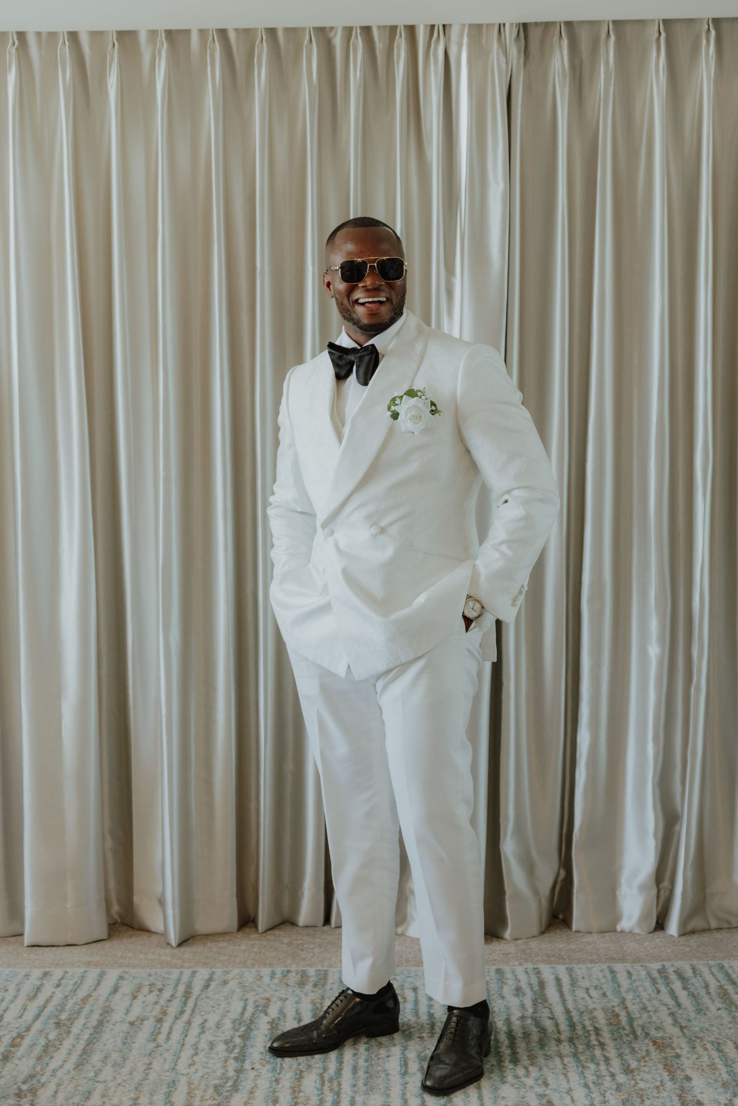 Stylish groom in white double-breasted suit with bow tie and sunglasses, smiling confidently indoors.
