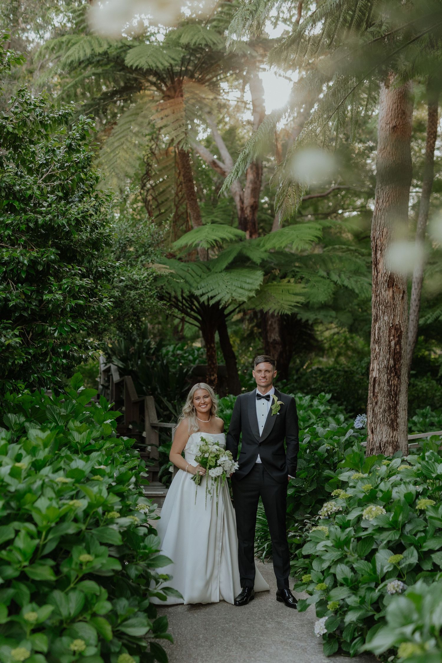Bride and groom pose on lush garden path surrounded by tropical ferns and green foliage.