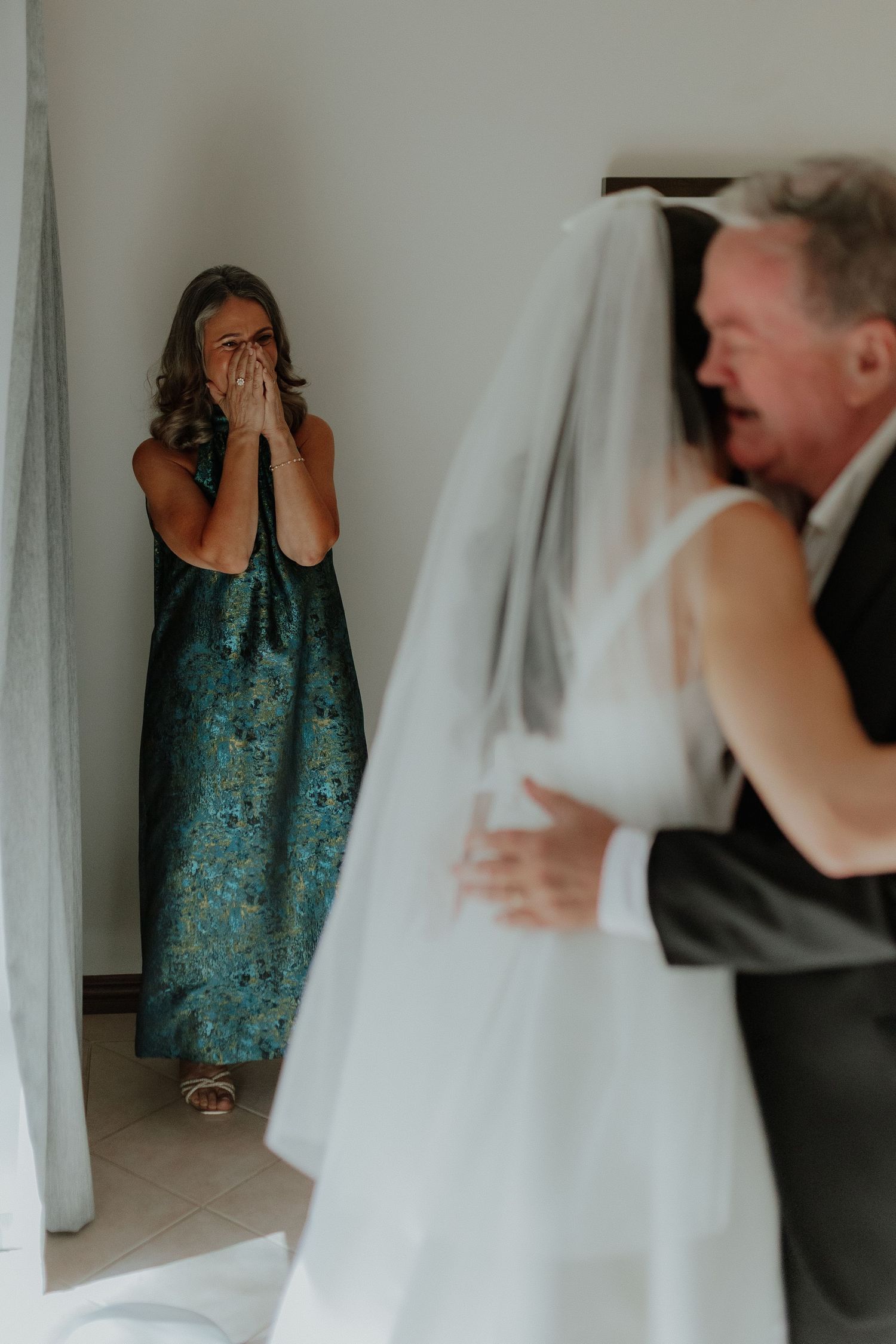 Emotional mother covers her face in tears watching bride and father share first look moment indoors.