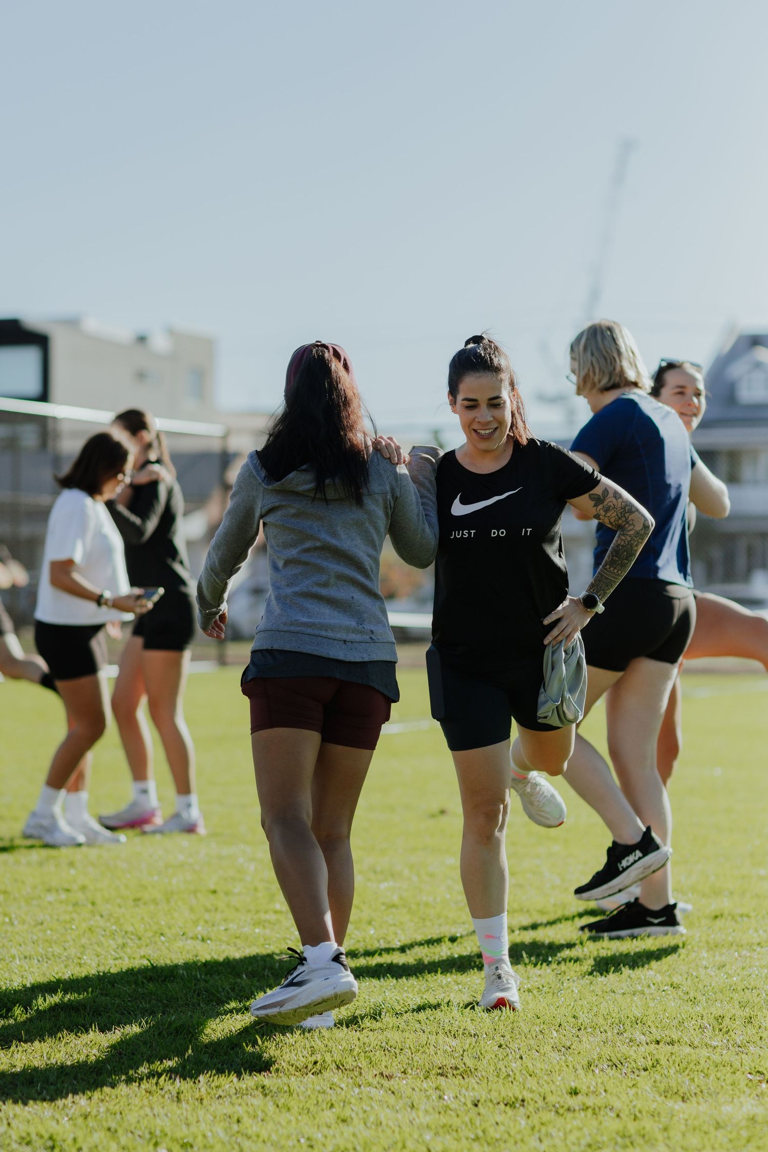 Two women laughing and walking together on a sunny outdoor sports field during a fitness event.