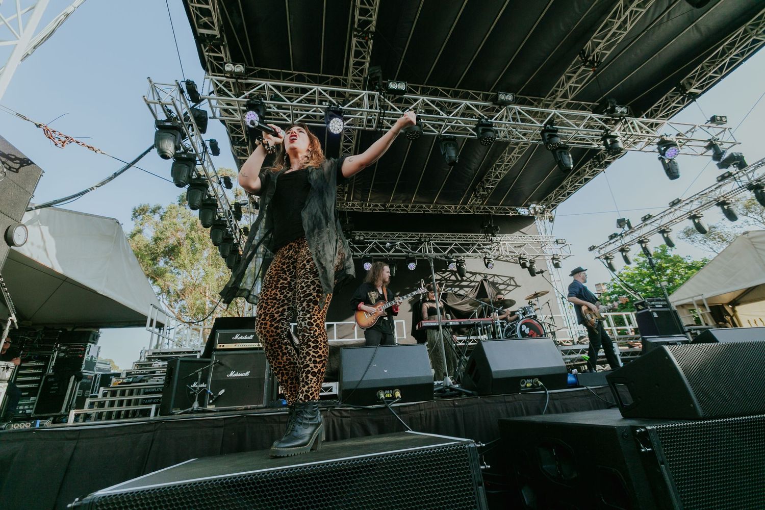 Female singer performing on outdoor festival stage wearing leopard print pants with band and speakers visible.