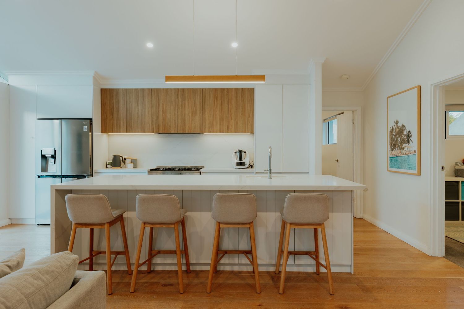 Modern open-plan kitchen with timber cabinetry, white island bench, bar stools, and stainless steel appliances.