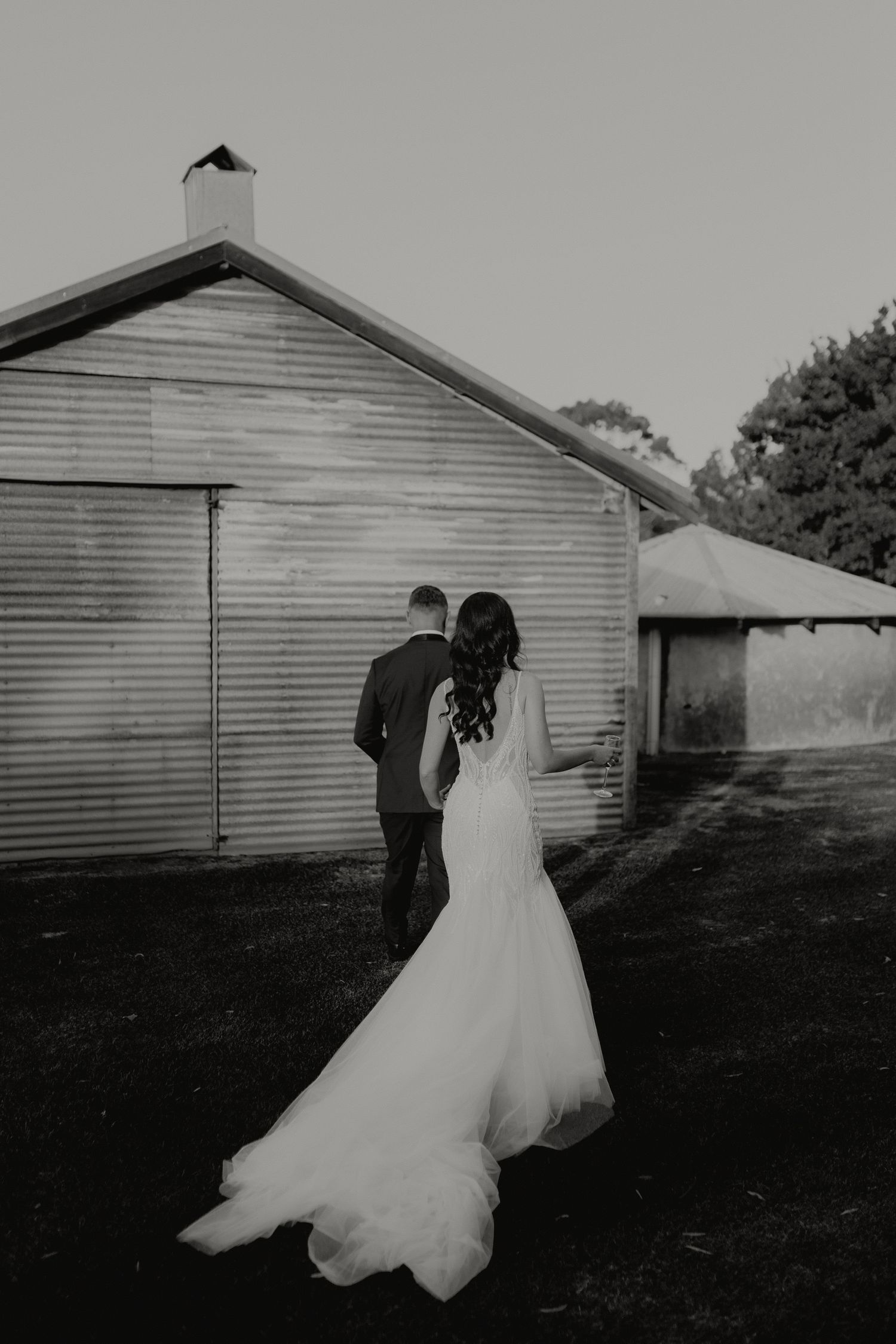 Black and white photo of bride and groom walking toward rustic barn in wedding attire.