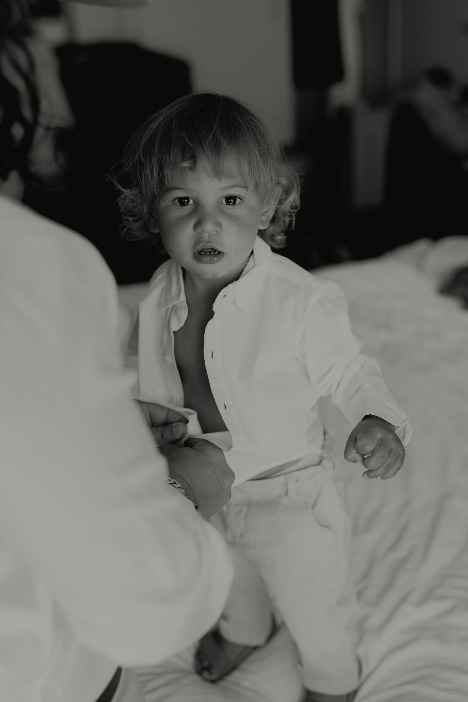 Adorable toddler in white linen outfit sitting on bed in black and white wedding photo.
