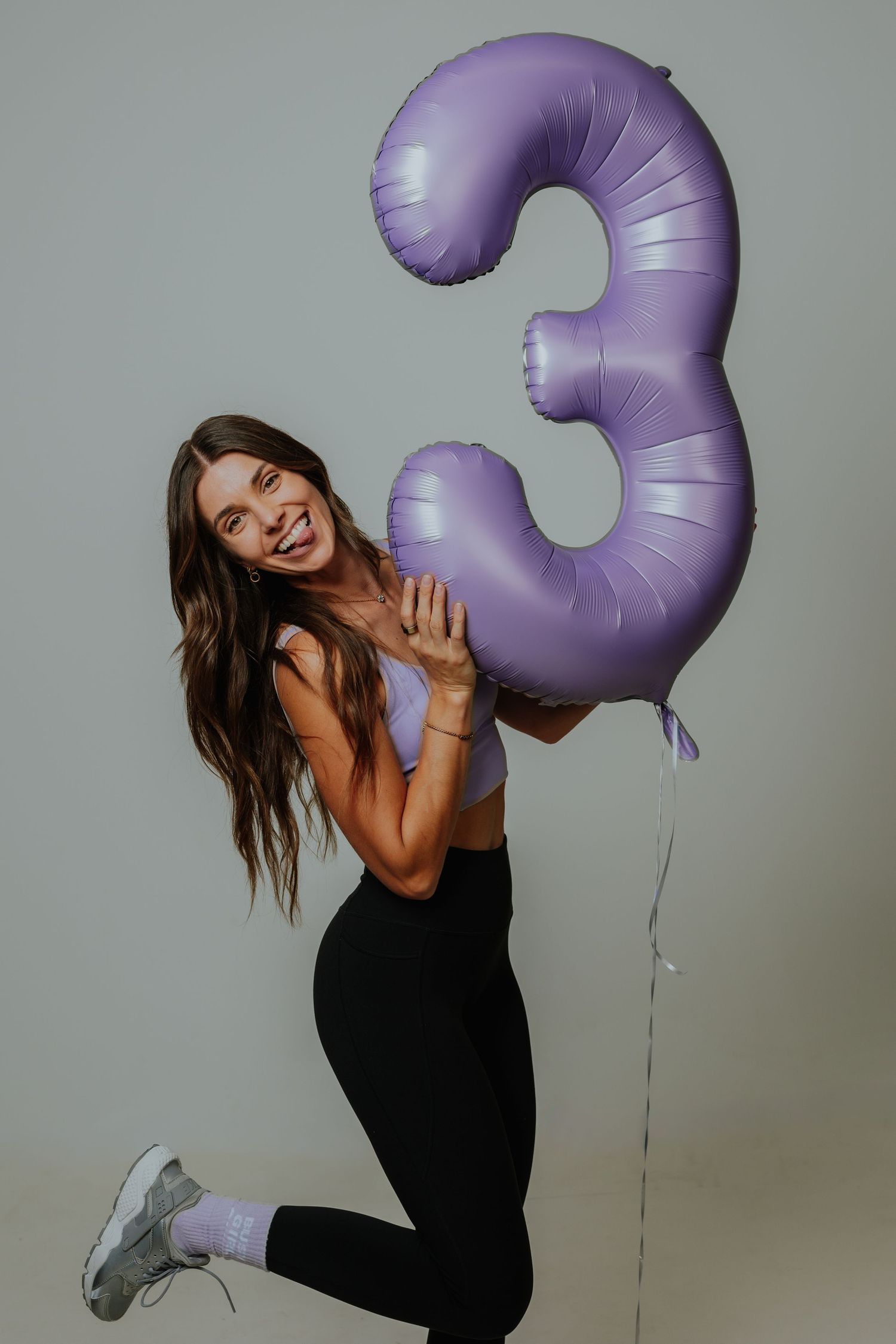 Happy woman in lavender crop top holding a purple number 3 balloon, jumping playfully.