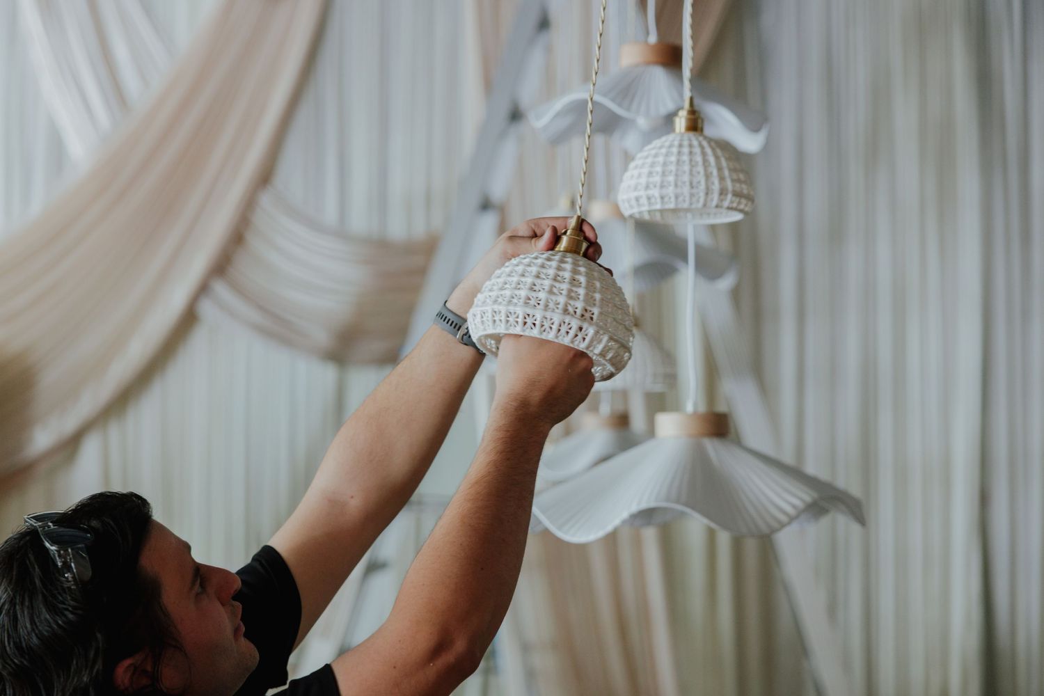 Person adjusting white ceramic pendant lights against elegant draped white fabric curtains.