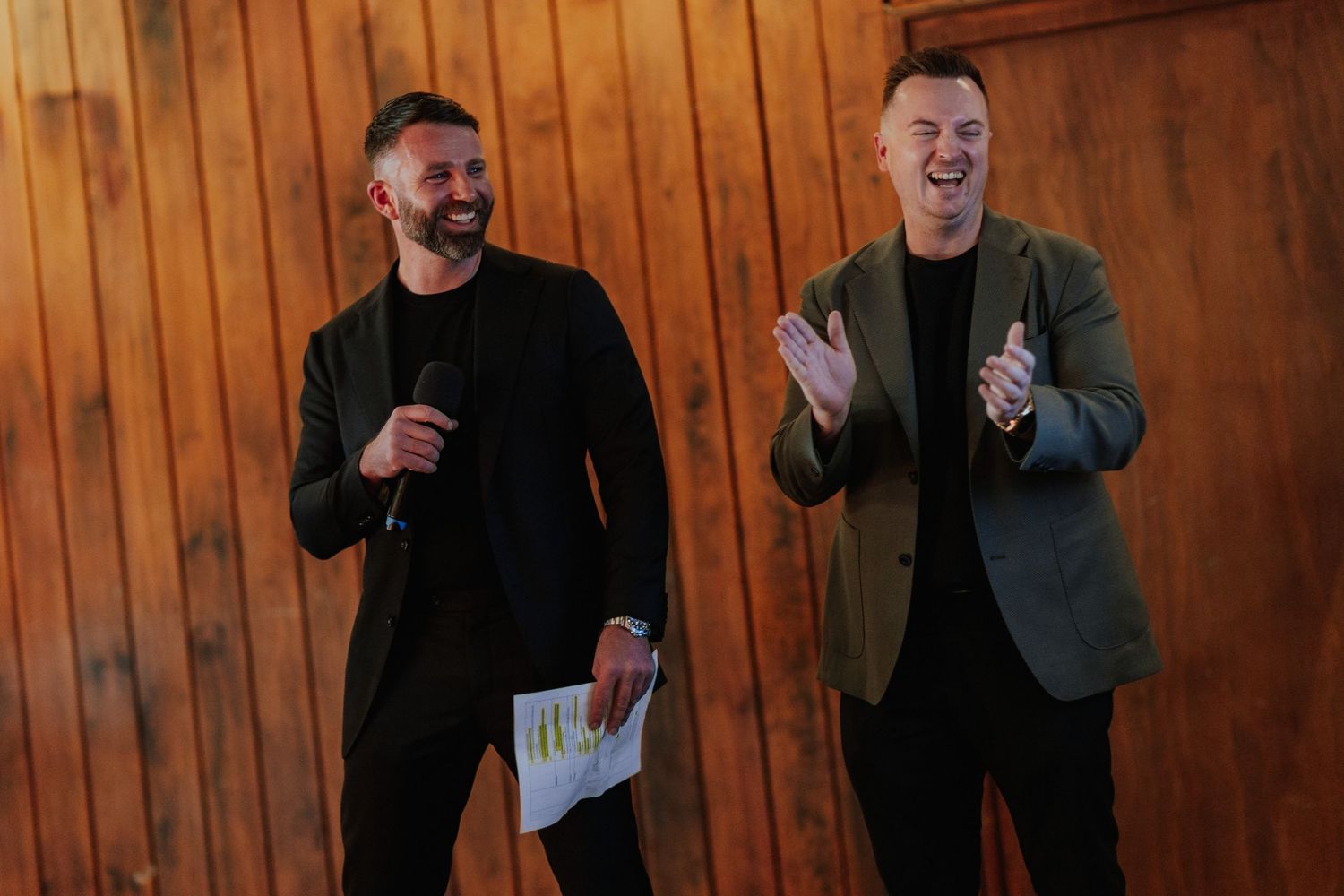 Two men in dark blazers smiling and presenting at a formal event against a wooden backdrop.