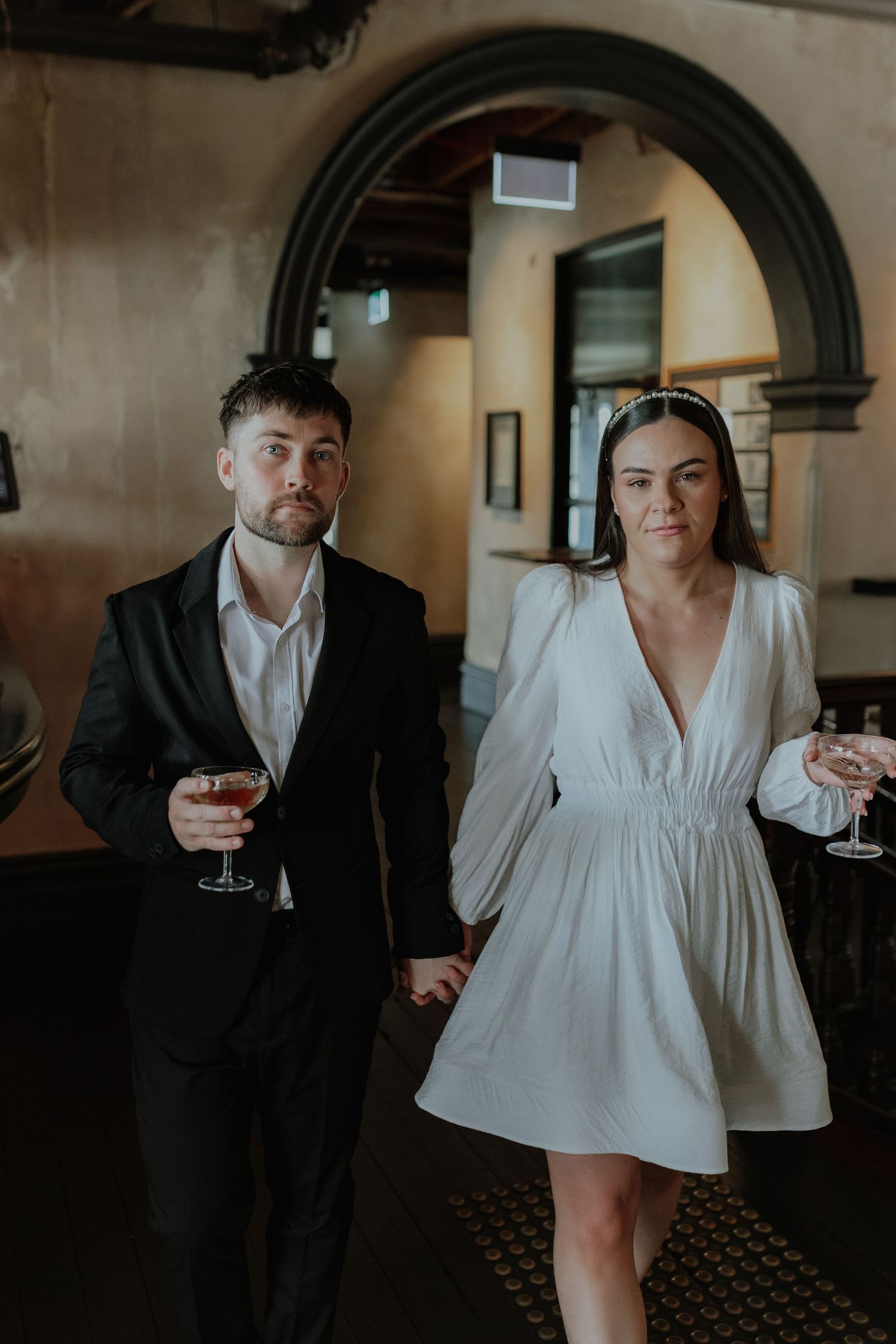 Stylish couple holding cocktails walking through an elegant venue, woman in white mini dress.