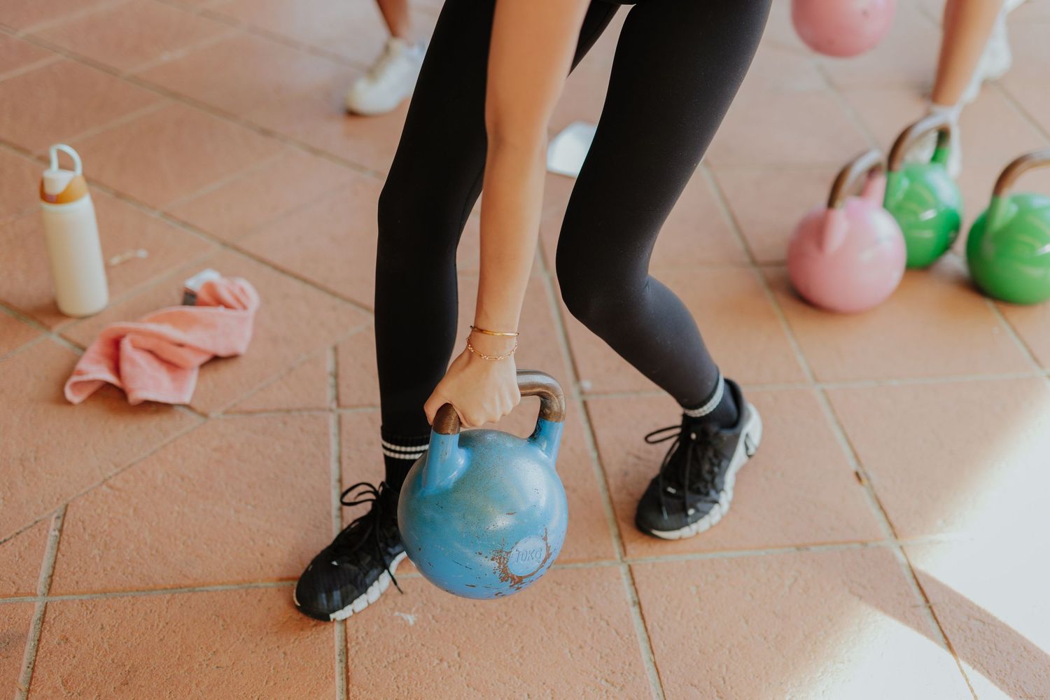 Woman lifting a blue kettlebell during an indoor workout session on tiled floor.