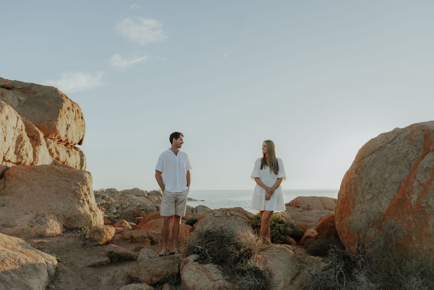 Couple in white outfits facing each other on rocky beach with calm ocean in the background.