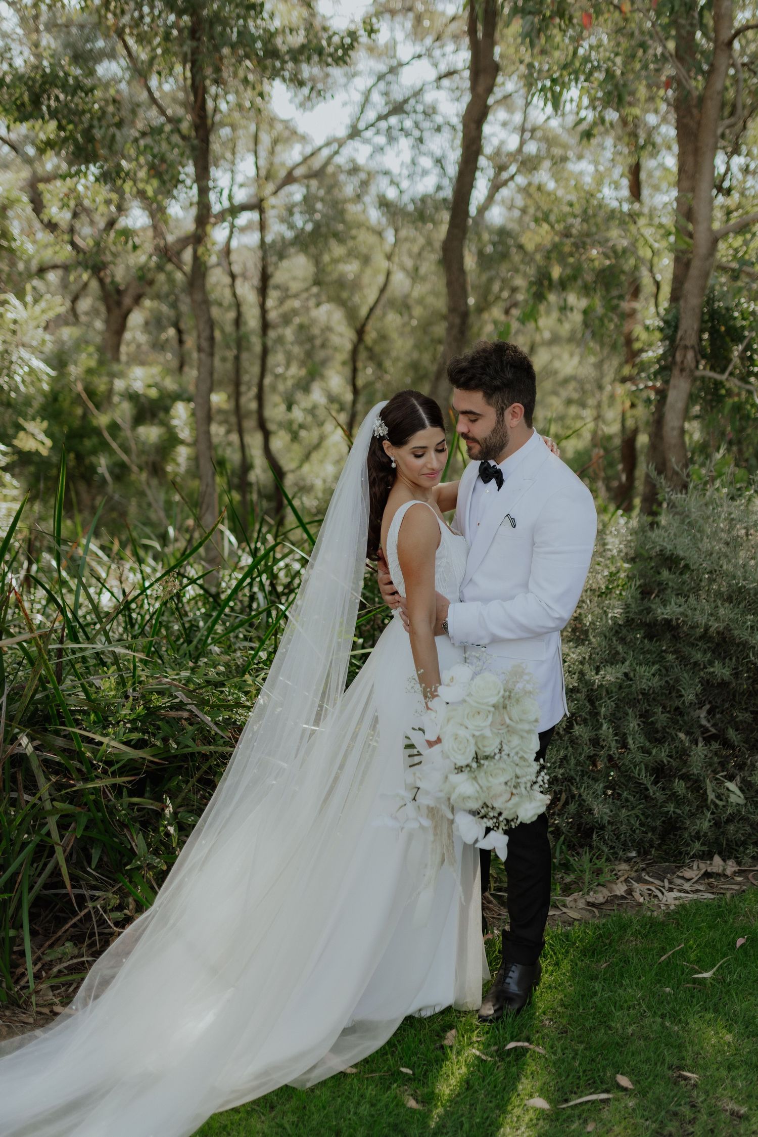 Bride in white gown with long veil holds white bouquet as groom in white jacket embraces her outdoors.