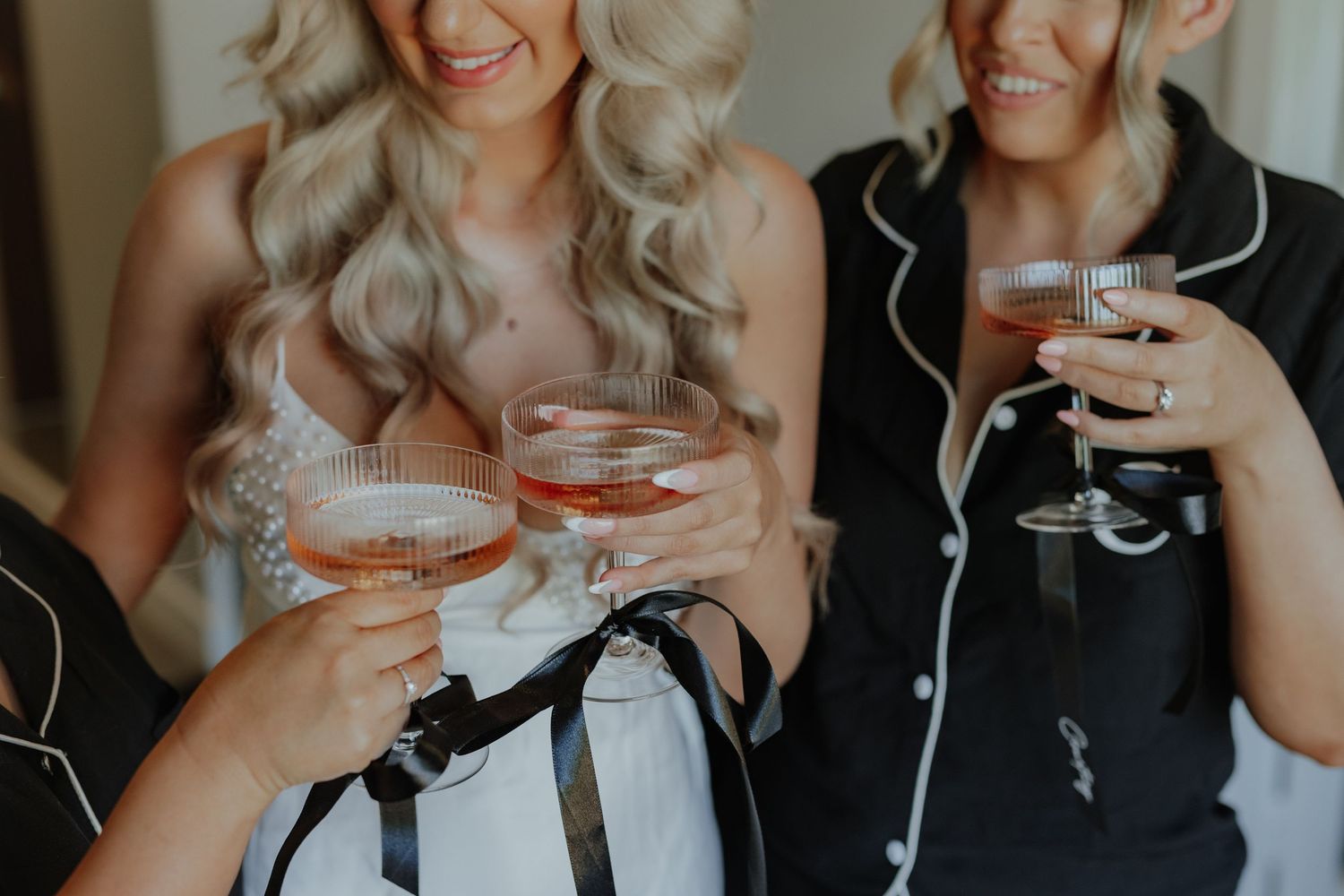 Bridesmaids in black satin pajamas toasting with pink cocktails during wedding preparations.