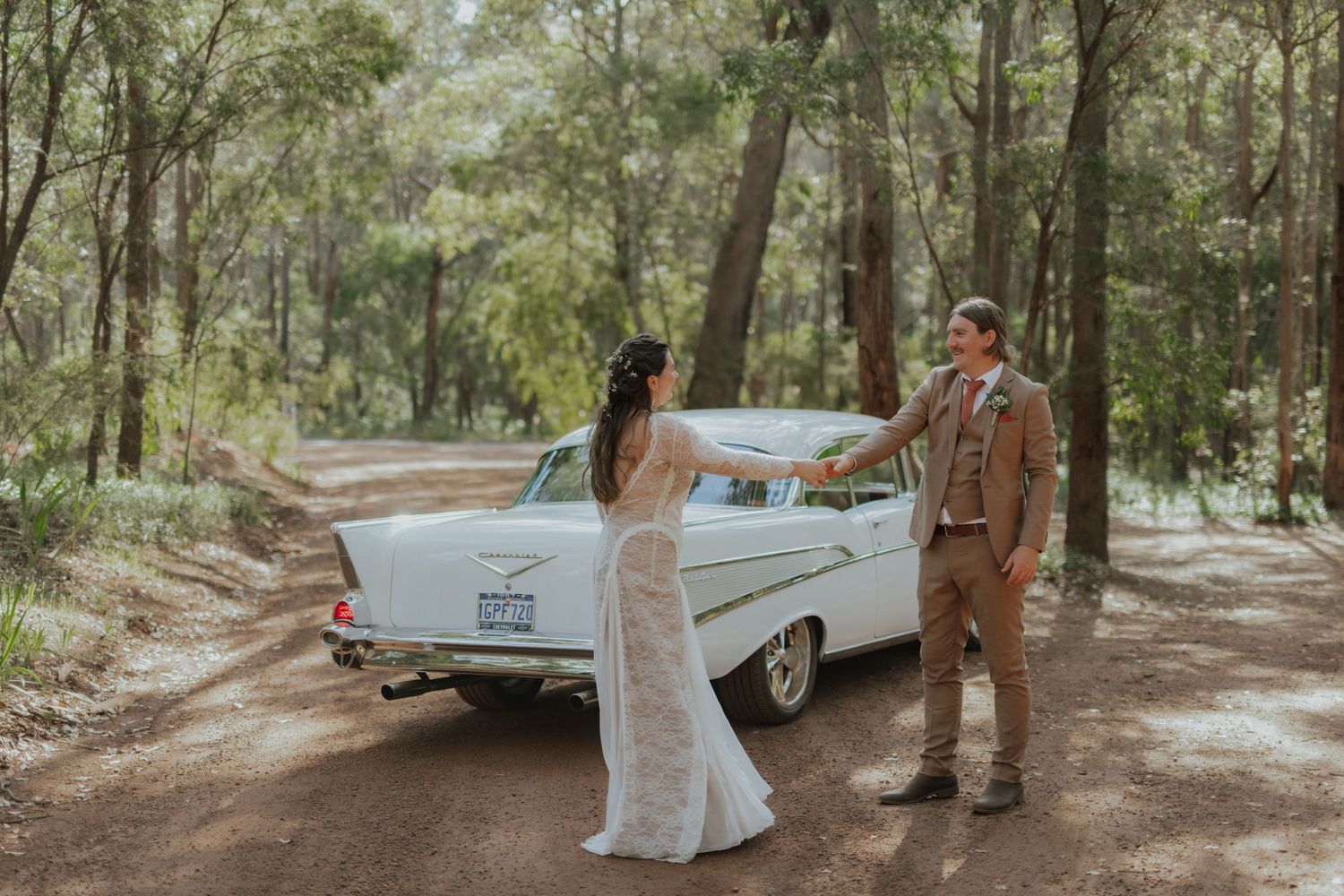 Bride and groom hold hands beside a vintage white classic car on a forest dirt road.
