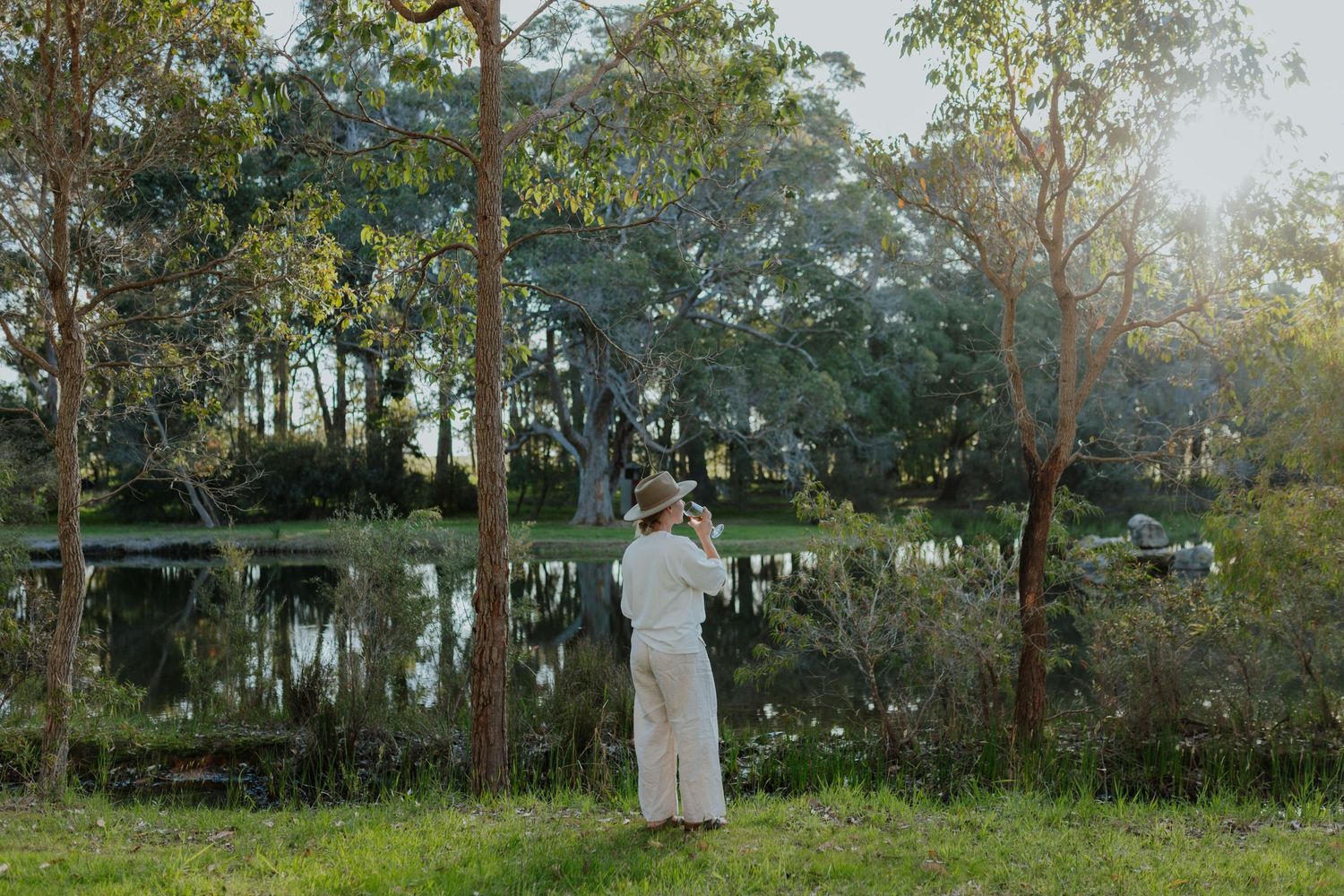 Person photographing a serene lake surrounded by lush trees in golden light.
