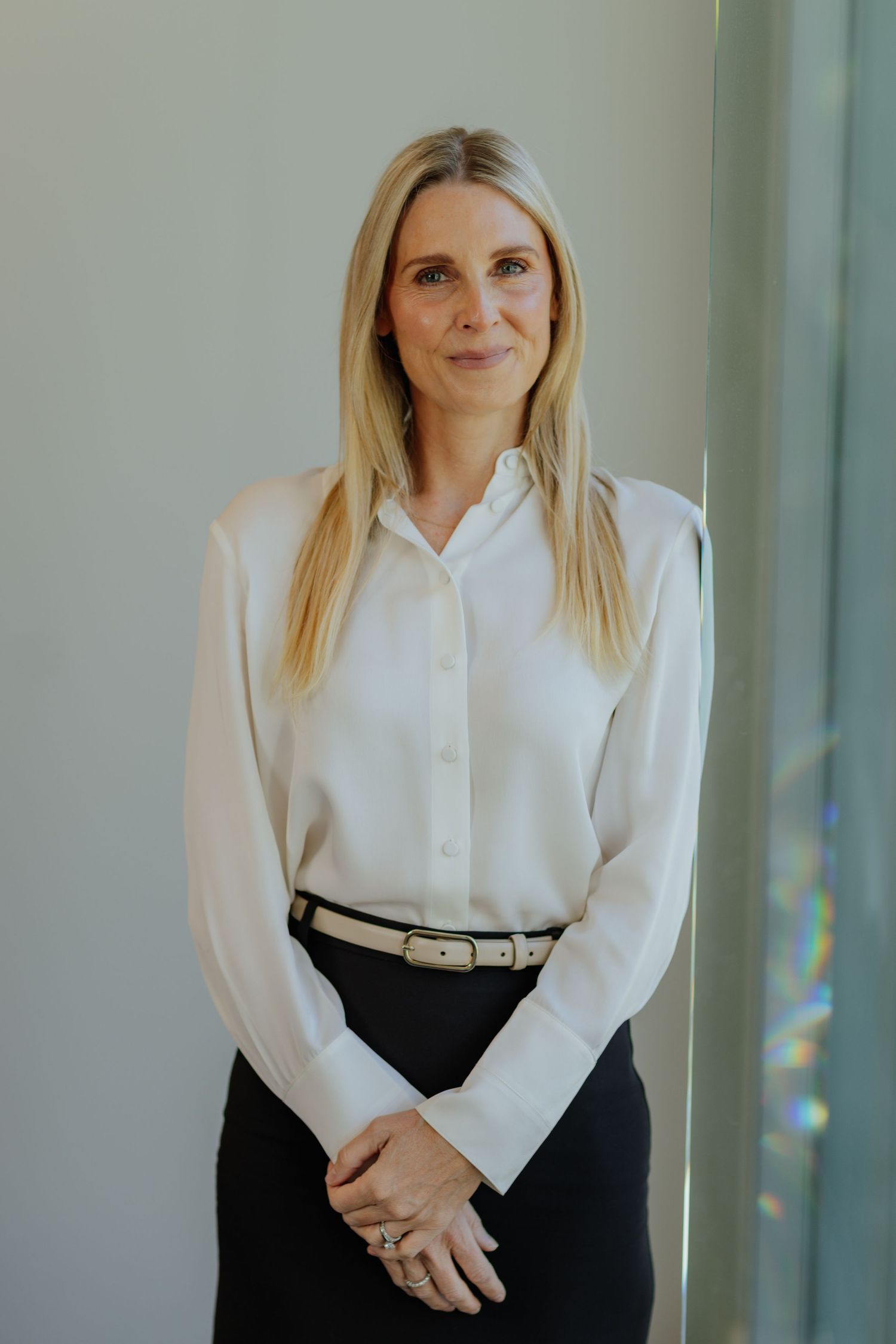 Professional blonde woman in white silk blouse and black skirt standing in modern office.