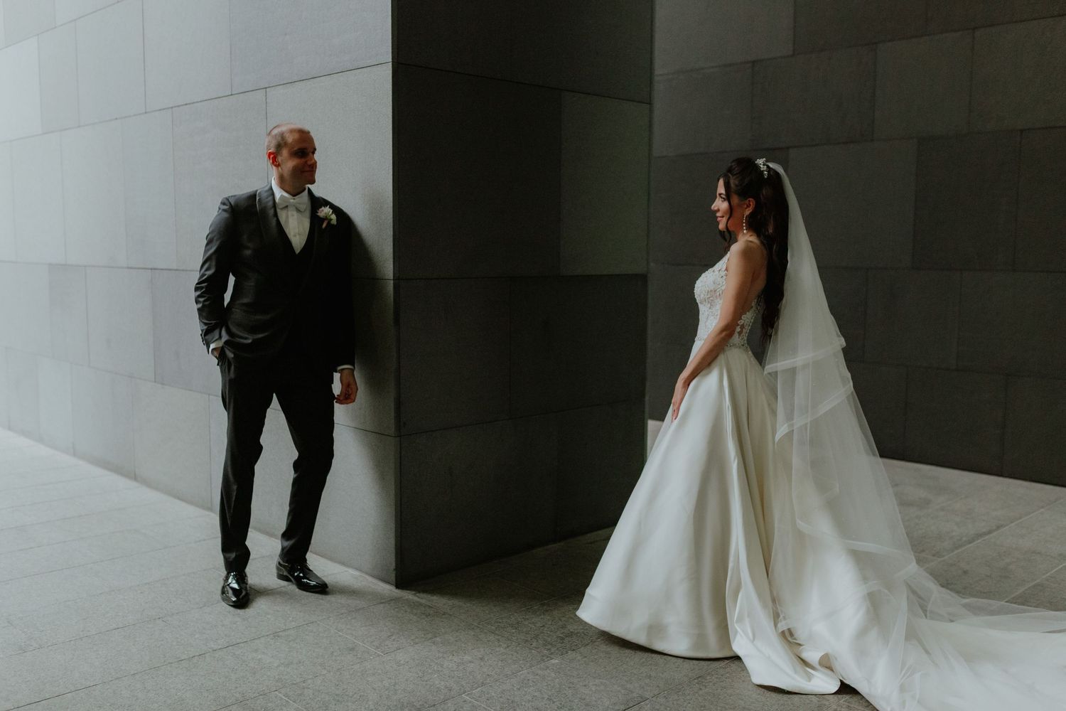 Bride in white gown with veil and groom in black suit posing against a modern stone wall.