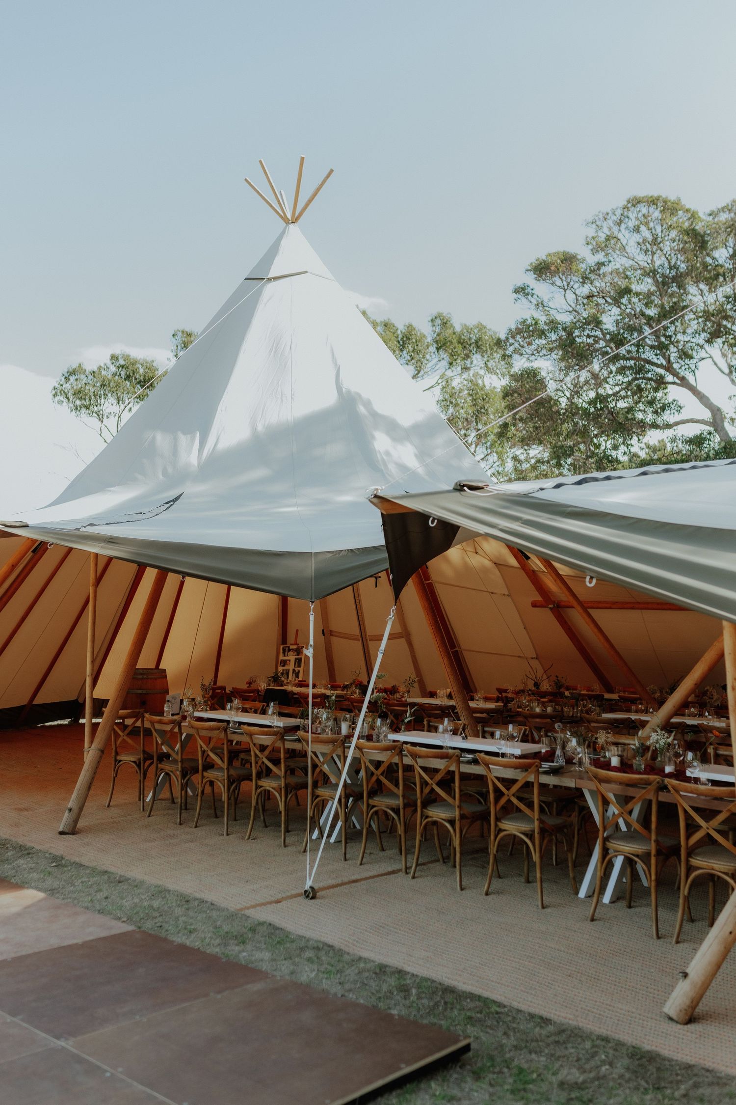 Elegant tipi wedding reception tent with long dining tables and chairs set up outdoors during daytime.