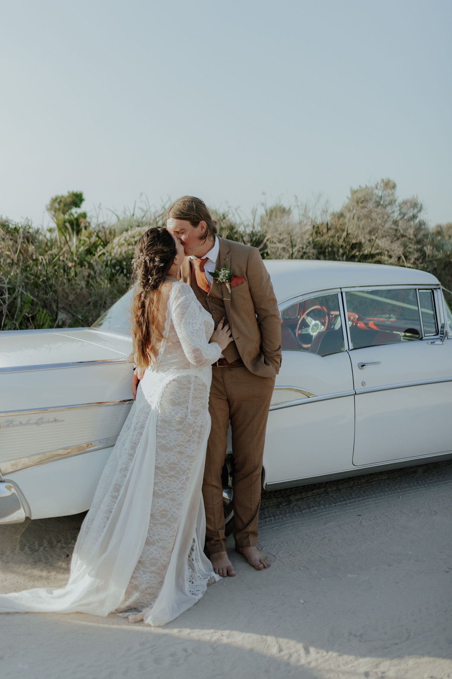 Bride and groom share a kiss leaning against vintage white classic car on sandy beach.