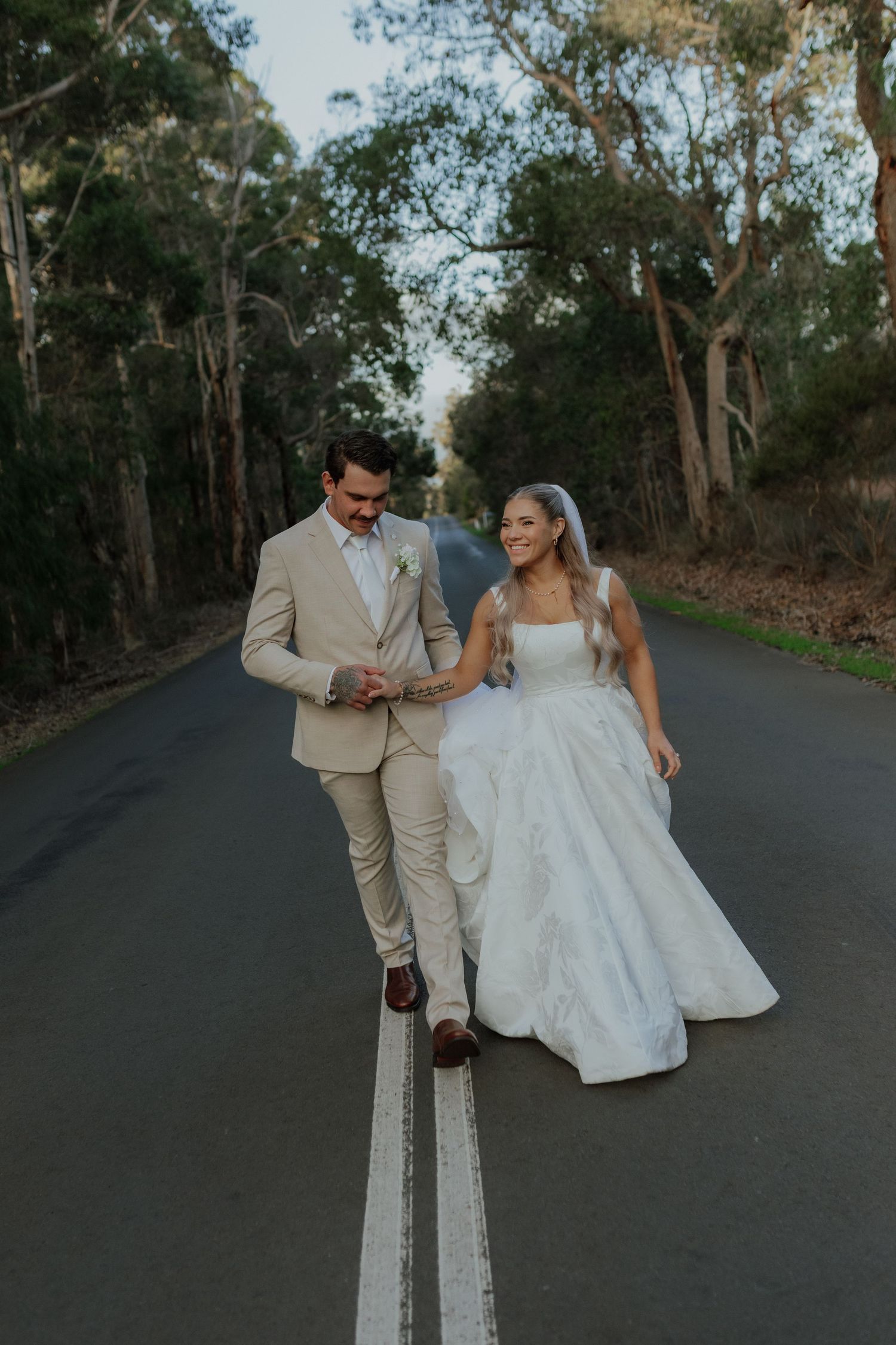 Bride and groom walking down tree-lined road, groom in beige suit and bride in ballgown.