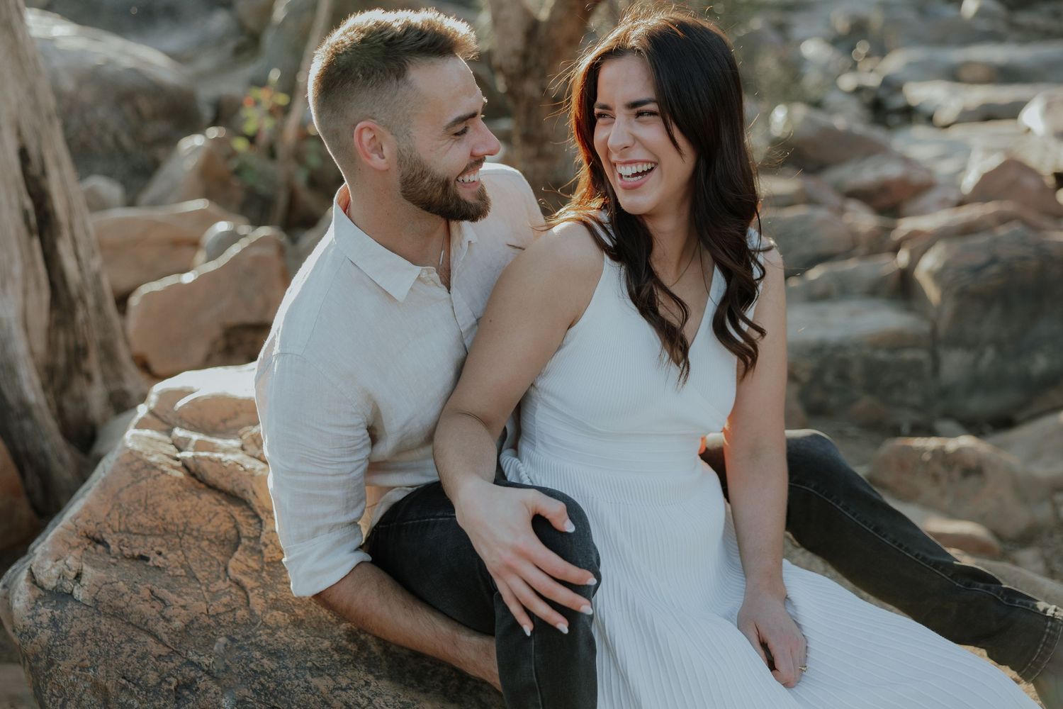 Joyful couple sitting on rocks outdoors laughing together in warm golden hour sunlight.
