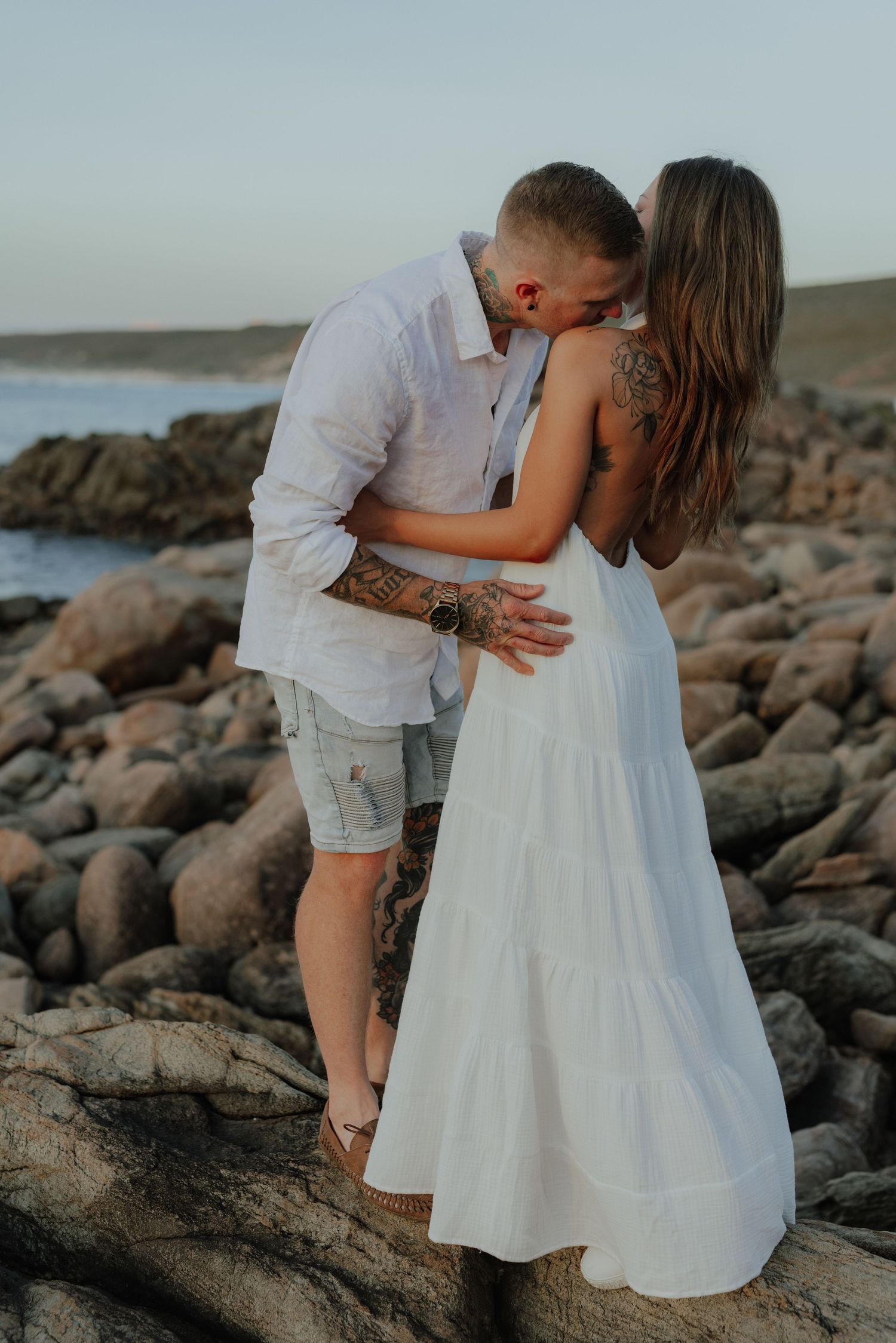 Couple embracing on rocky beach at sunset, man kissing woman in elegant white backless dress.