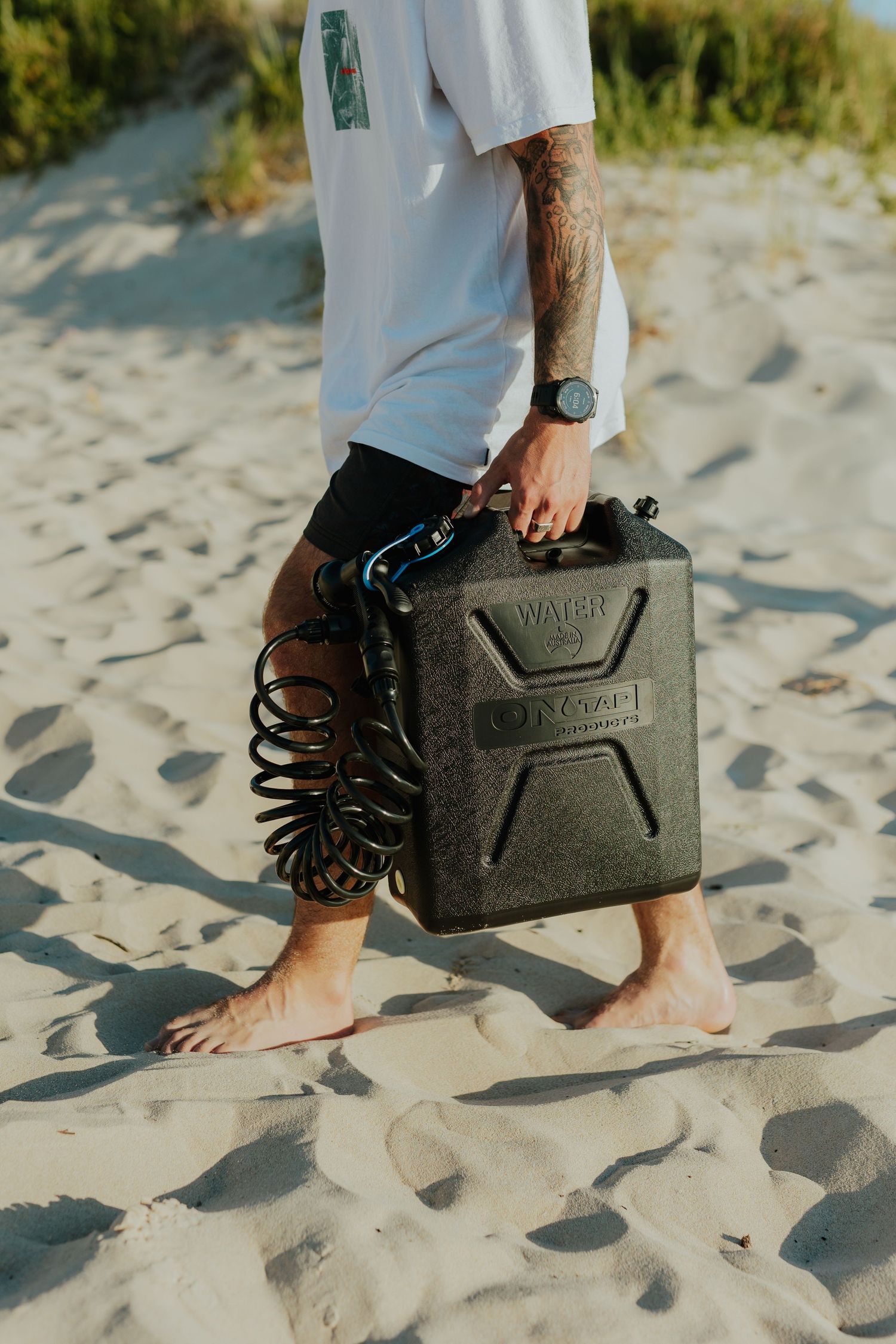 Tattooed man carrying a dark green water jerry can across sandy beach.