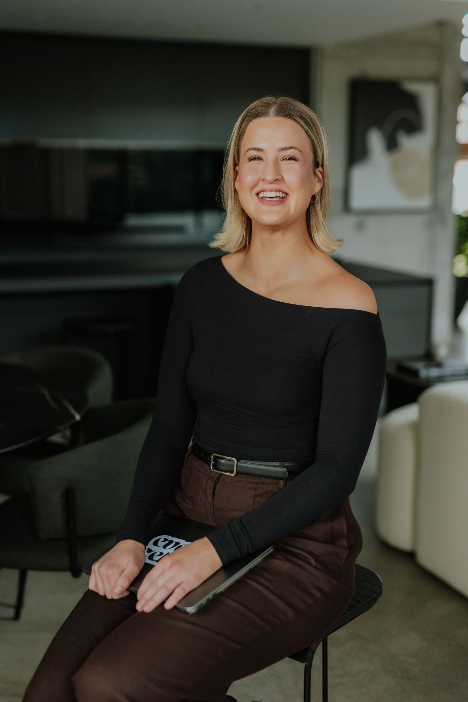 Smiling blonde woman in black off-shoulder top and brown pants sitting on a stool in a modern interior.