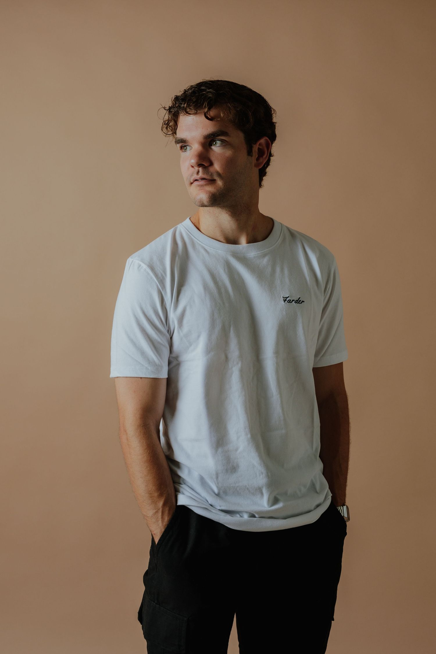 Young man wearing a white Farder branded t-shirt with black pants posed against a neutral beige background.