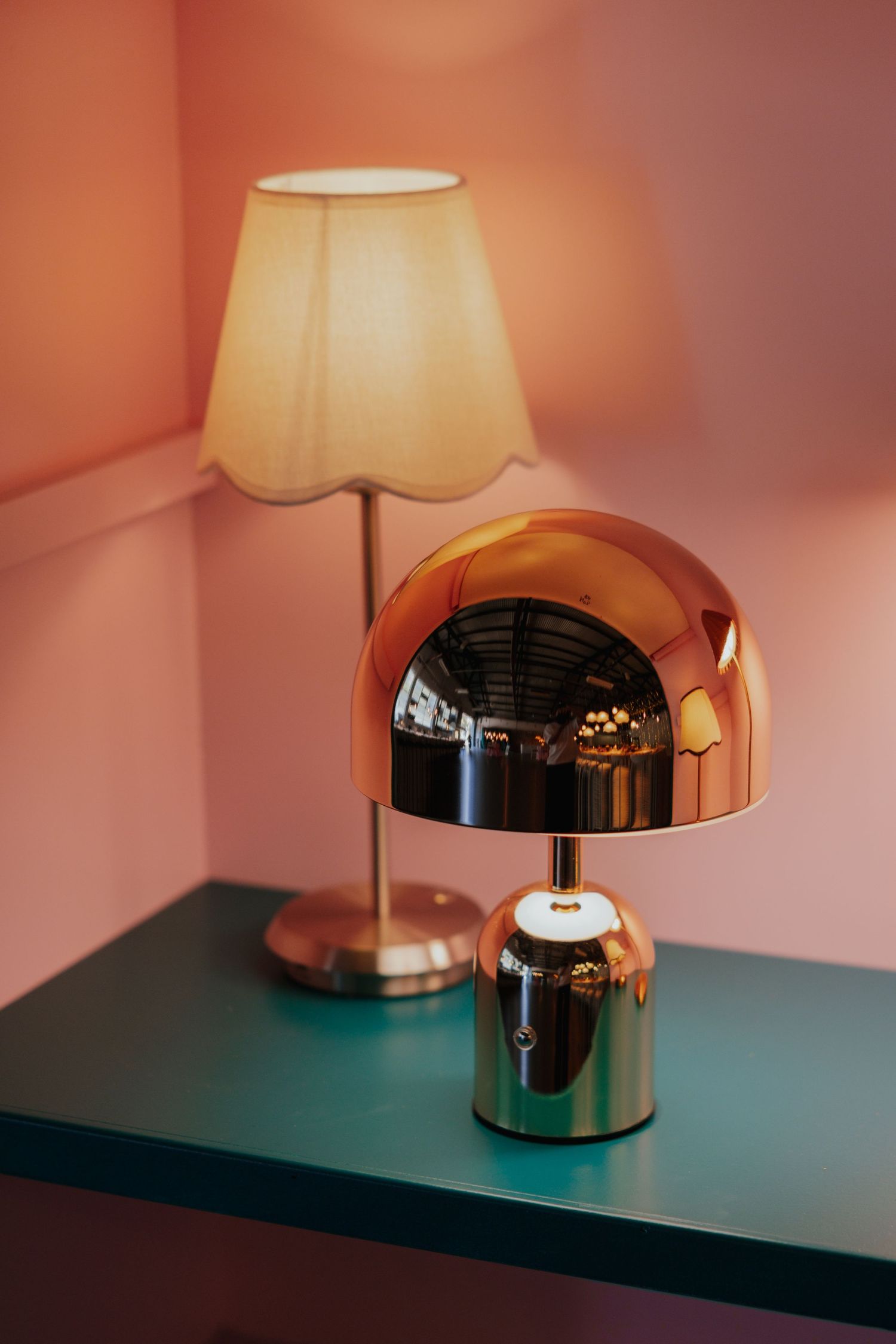 Rose gold dome lamp and cream scalloped shade table lamp on teal shelf in pink room.