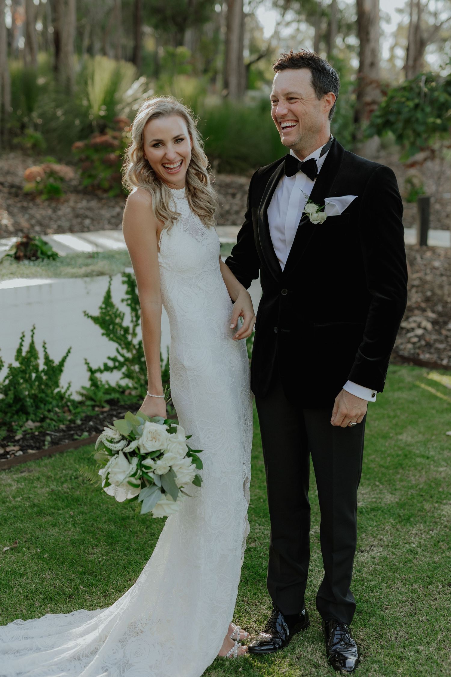 Smiling bride in white halter gown holding bouquet with groom in black velvet tuxedo outdoors.