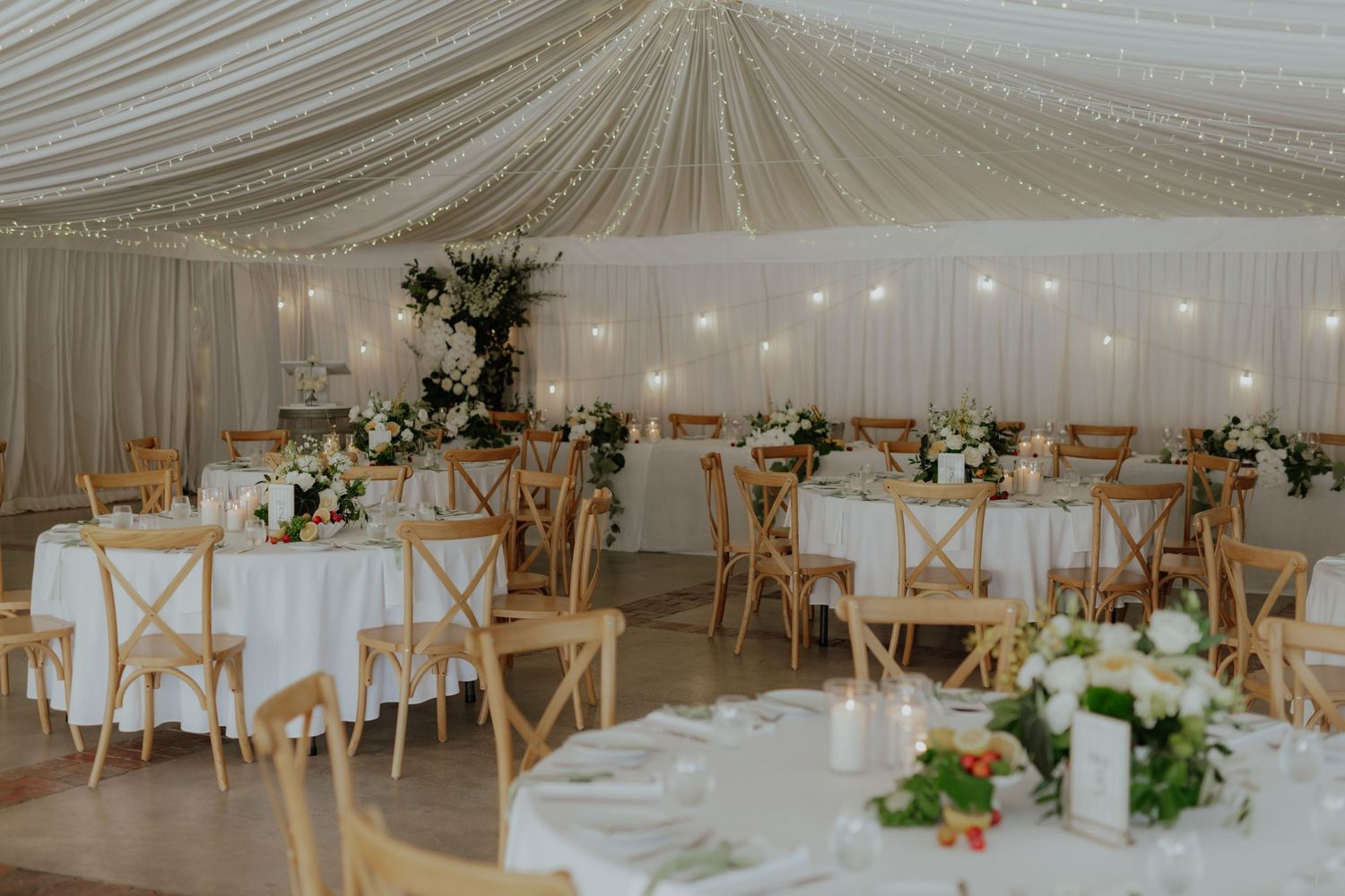 Elegant white marquee wedding reception with draped ceiling, wooden cross-back chairs, and floral centerpieces.